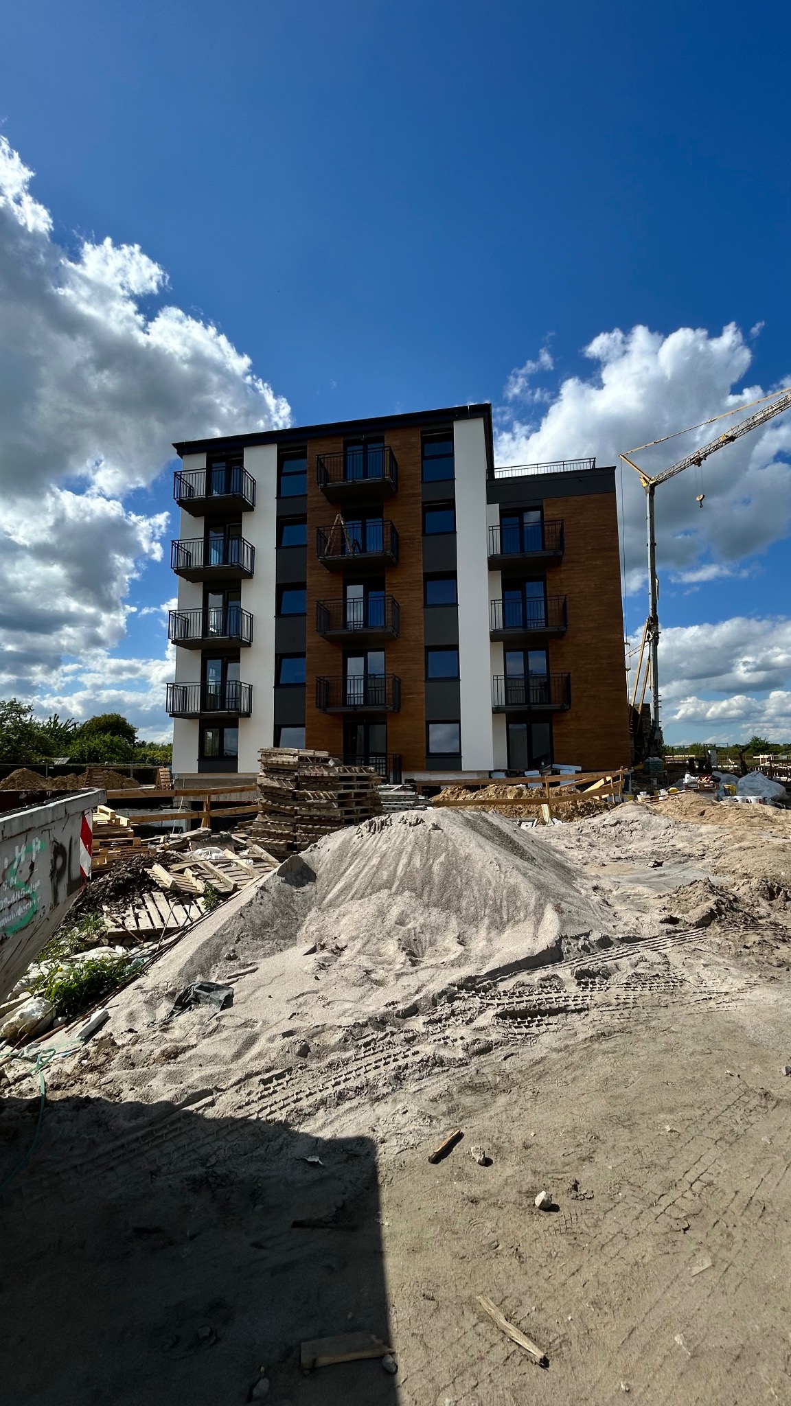 Wielopiętrowy budynek z balkonami w trakcie budowy, widoczny żuraw budowlany. Hałda piasku i materiały budowlane na pierwszym planie. Błękitne niebo z chmurami w tle.