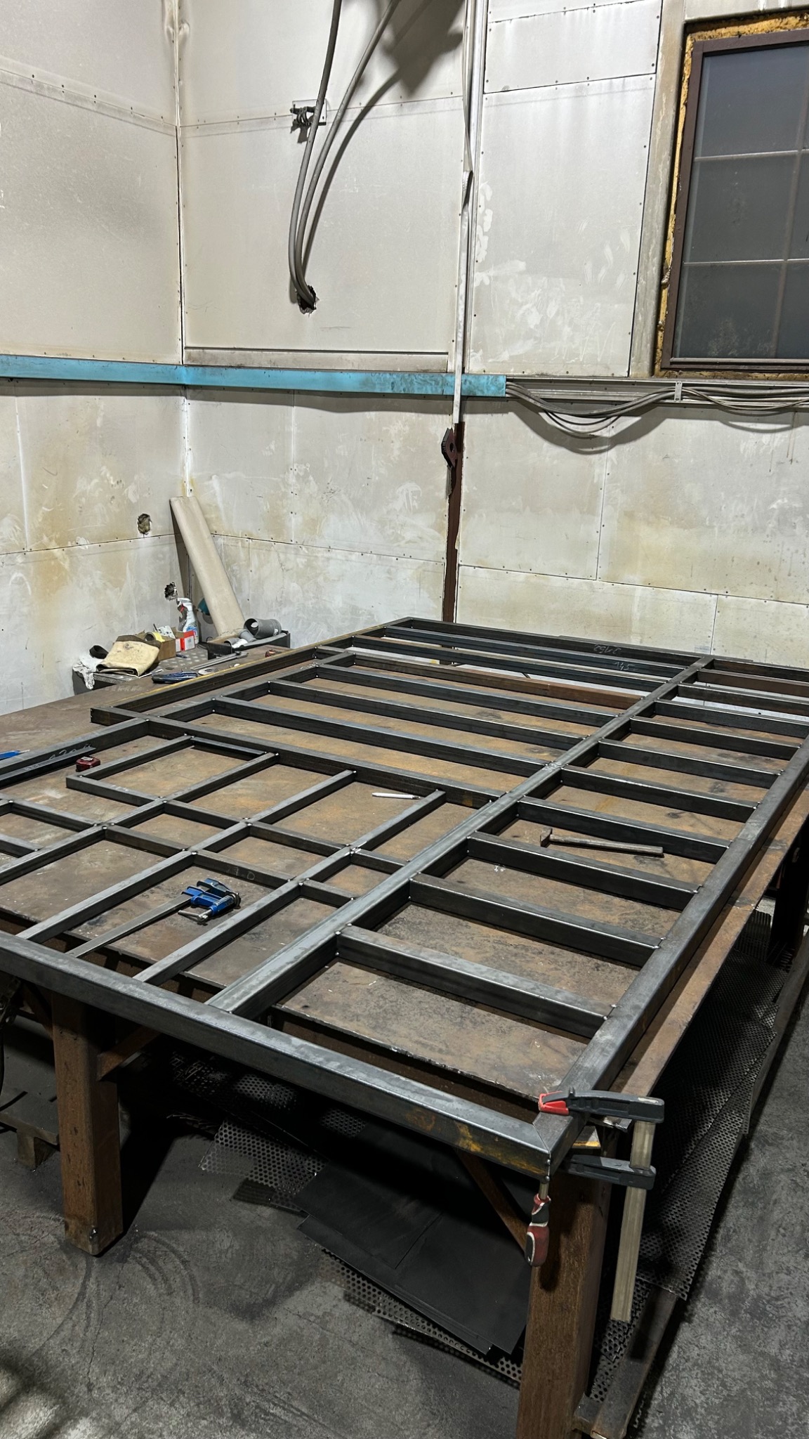 Stalowa konstrukcja kratowa na stole warsztatowym, z widocznymi śladami spawania i zaciskami. W tle ściana warsztatu i okno.