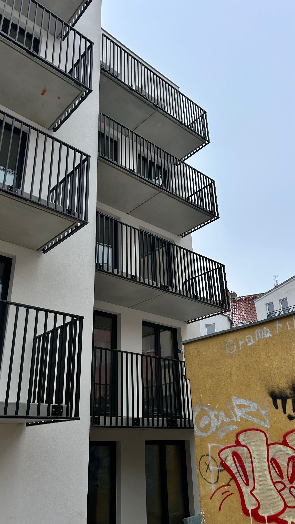 Biały budynek z balkonami o czarnych, metalowych balustradach. Ujęcie z dołu, widoczne graffiti na sąsiedniej ścianie. Minimalistyczny design.
