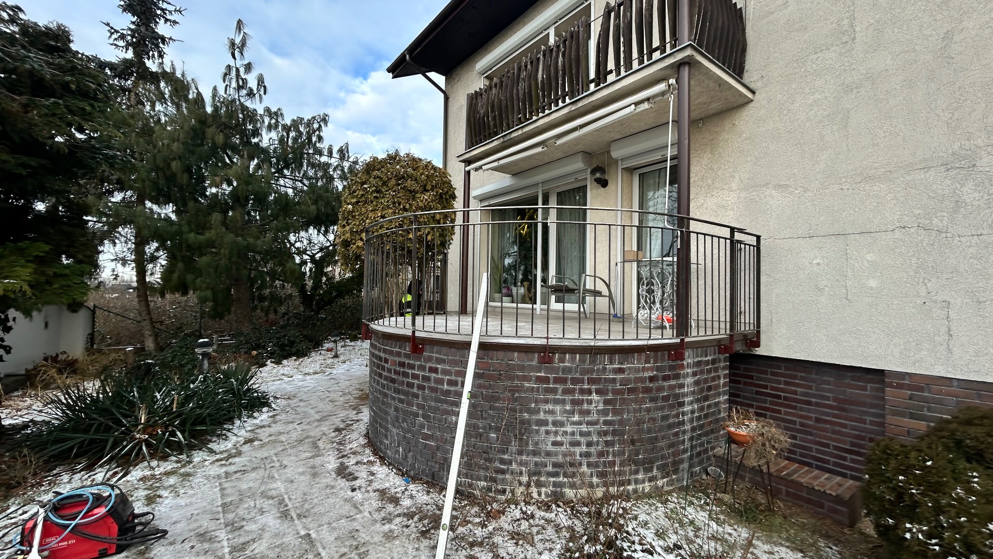 Balkon z metalową balustradą o zaokrąglonym kształcie, wsparty na ceglanej konstrukcji. Na balkonie widoczne krzesła i stolik. Na pierwszym planie poziomica i spawarka.