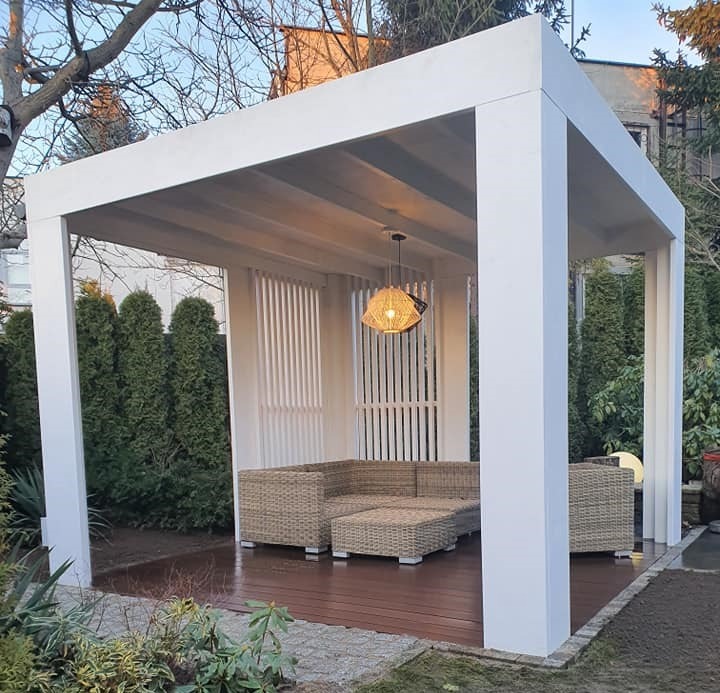 Biała, nowoczesna konstrukcja z drewnianą podłogą i rattanowymi meblami ogrodowymi, oświetlona wiszącą lampą z naturalnego materiału, otoczona zielenią.