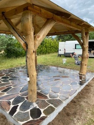 Niedokończona altana z drewnianą konstrukcją i kamienną posadzką z nieregularnych płyt, w tle biały bus i pracownik w trakcie pracy.