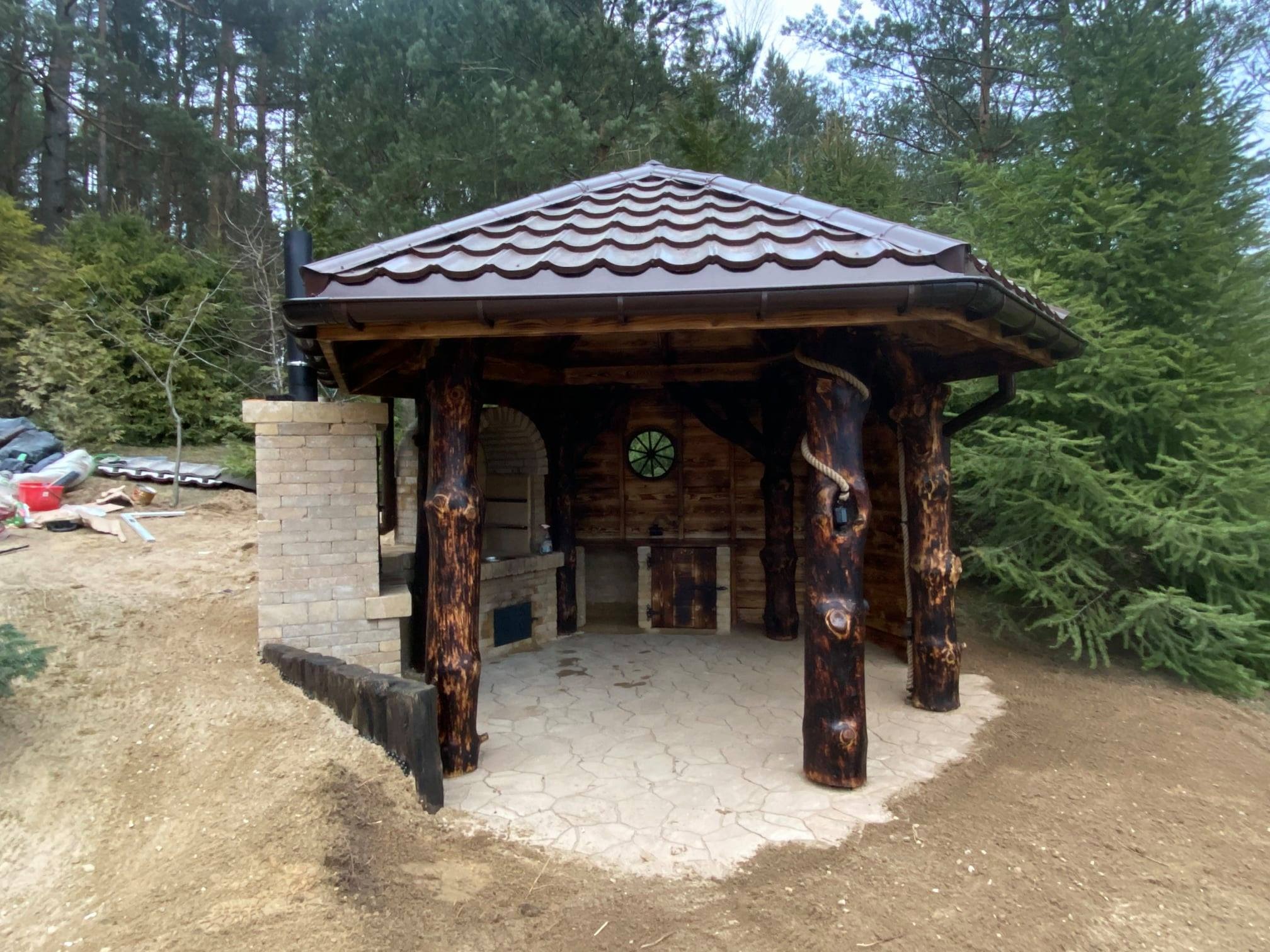 Rustykalna altana z drewnianymi kolumnami imitującymi pnie drzew, murowanym grillem i piecem, brązowym dachem z blachodachówki i podłogą z płyt kamiennych na działce w trakcie zagospodarowywania.