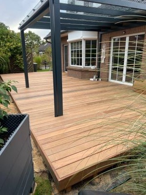 Nowoczesny taras drewniany z pergolą w kolorze antracytowym, widok na dom z cegły i zadbany ogród.