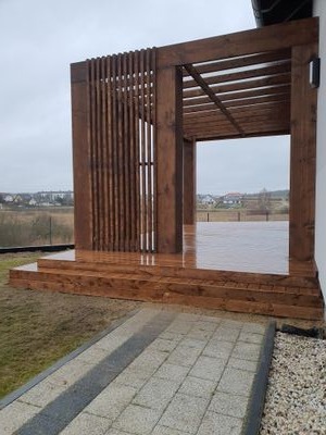 Nowoczesna, ciemnobrązowa konstrukcja drewniana z pionowymi listwami, prawdopodobnie pergola, na podwyższonym tarasie, widok na okolicę w tle, pochmurny dzień.