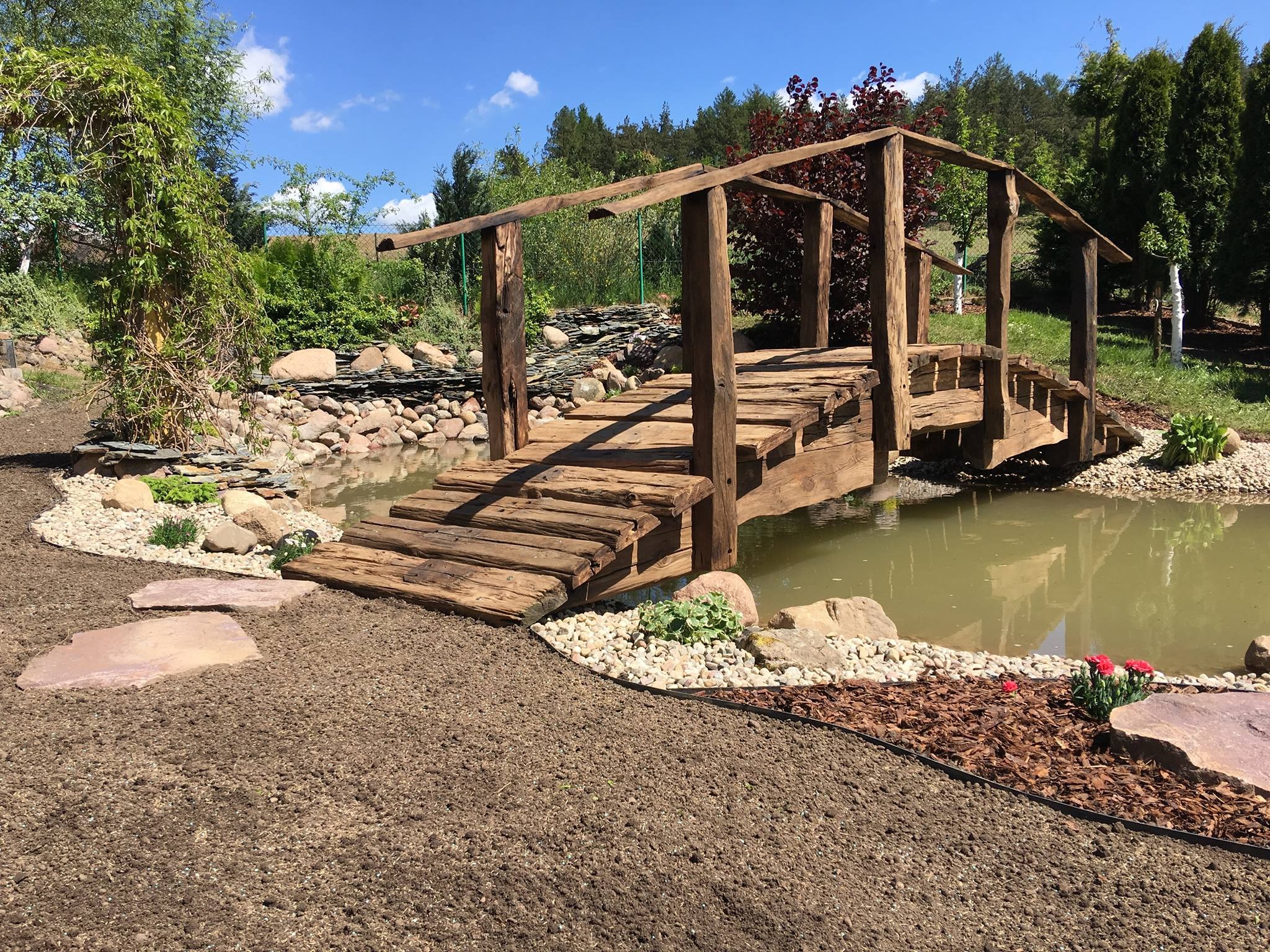 Rustykalny drewniany mostek nad stawem w ogrodzie, z elementami kamiennymi i roślinnością ozdobną, w słoneczny dzień.