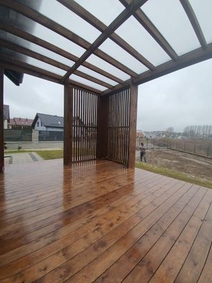 Zadaszony taras z drewnianą podłogą i pionowymi panelami, widok na posesję z domem w tle, lekko deszczowa pogoda.