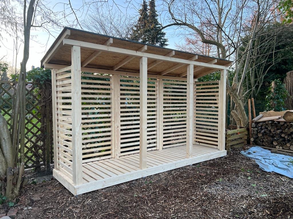 Surowa, drewniana konstrukcja zadaszonej przestrzeni z listwami, prawdopodobnie na drewno, w ogrodzie, z ułożonymi polanami w tle i rozłożoną plandeką.