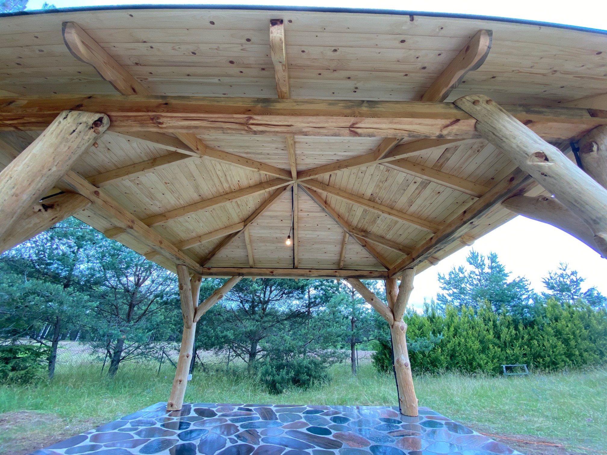 Wnętrze altany z drewnianą konstrukcją i kamienną posadzką, widok na zewnątrz z drzewami i zielenią.