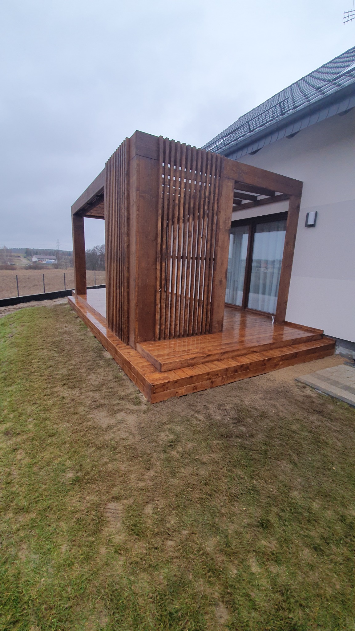Nowoczesna, drewniana konstrukcja z pionowymi lamelami, przylegająca do domu z widocznym fragmentem dachu i trawnikiem w tle.