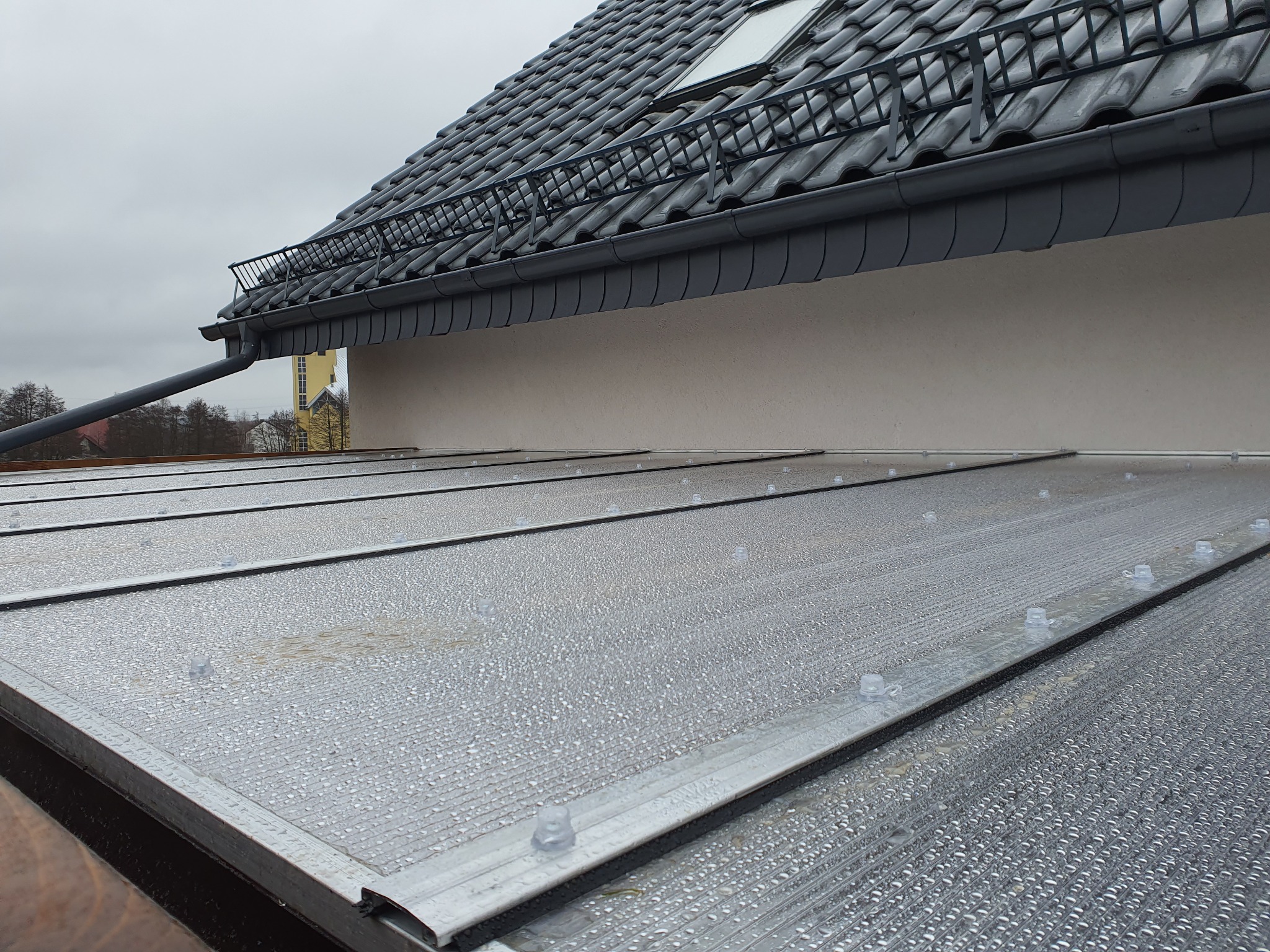 Przezroczyste panele z tworzywa sztucznego, prawdopodobnie poliwęglanu, tworzące zadaszenie tarasu lub werandy, widoczne krople wody, szare rynny i fragment dachu z dachówką w tle.