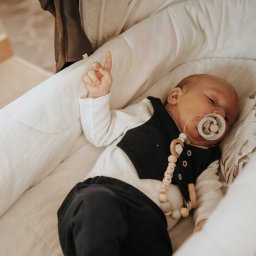 WK-FOTO Wojciech Kras Fotografia - Noworodek śpiący w białym, miękkim koszu Mojżesza z podniesioną rączką, ubrany w białe body, czarną kamizelkę i spodnie, z drewnianym smoczkiem z napisem 'MAKS'.