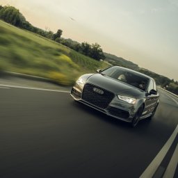WK-FOTO Wojciech Kras Fotografia - Dynamiczne ujęcie szarego Audi A3 na zakręcie drogi, z rozmytym tłem zielonych wzgórz i pochmurnego nieba, podkreślające prędkość i nowoczesny design auta.