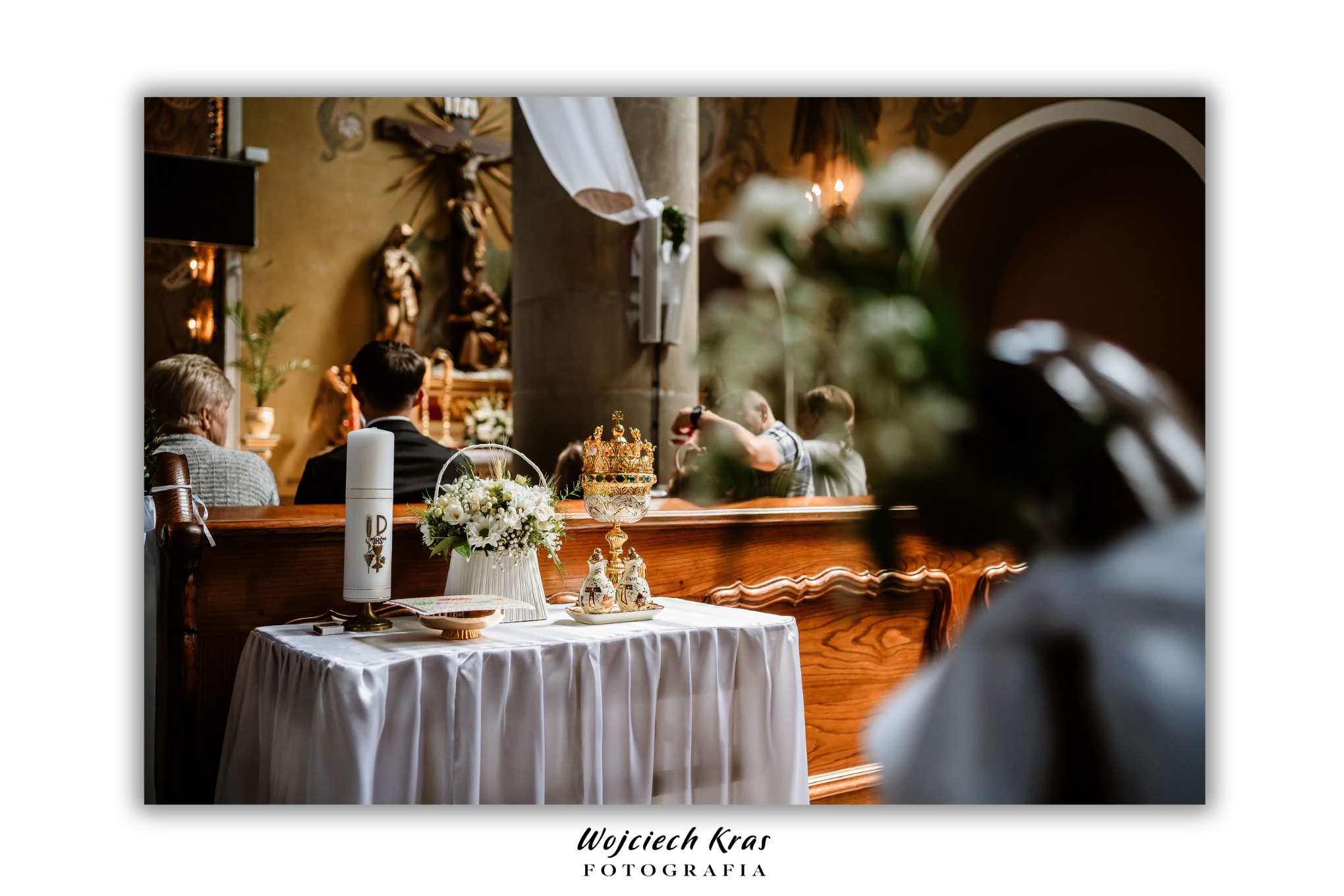 Dekoracja ołtarza z bukietem białych kwiatów w koszyku, świecą i kielichem podczas ceremonii ślubnej w kościele.