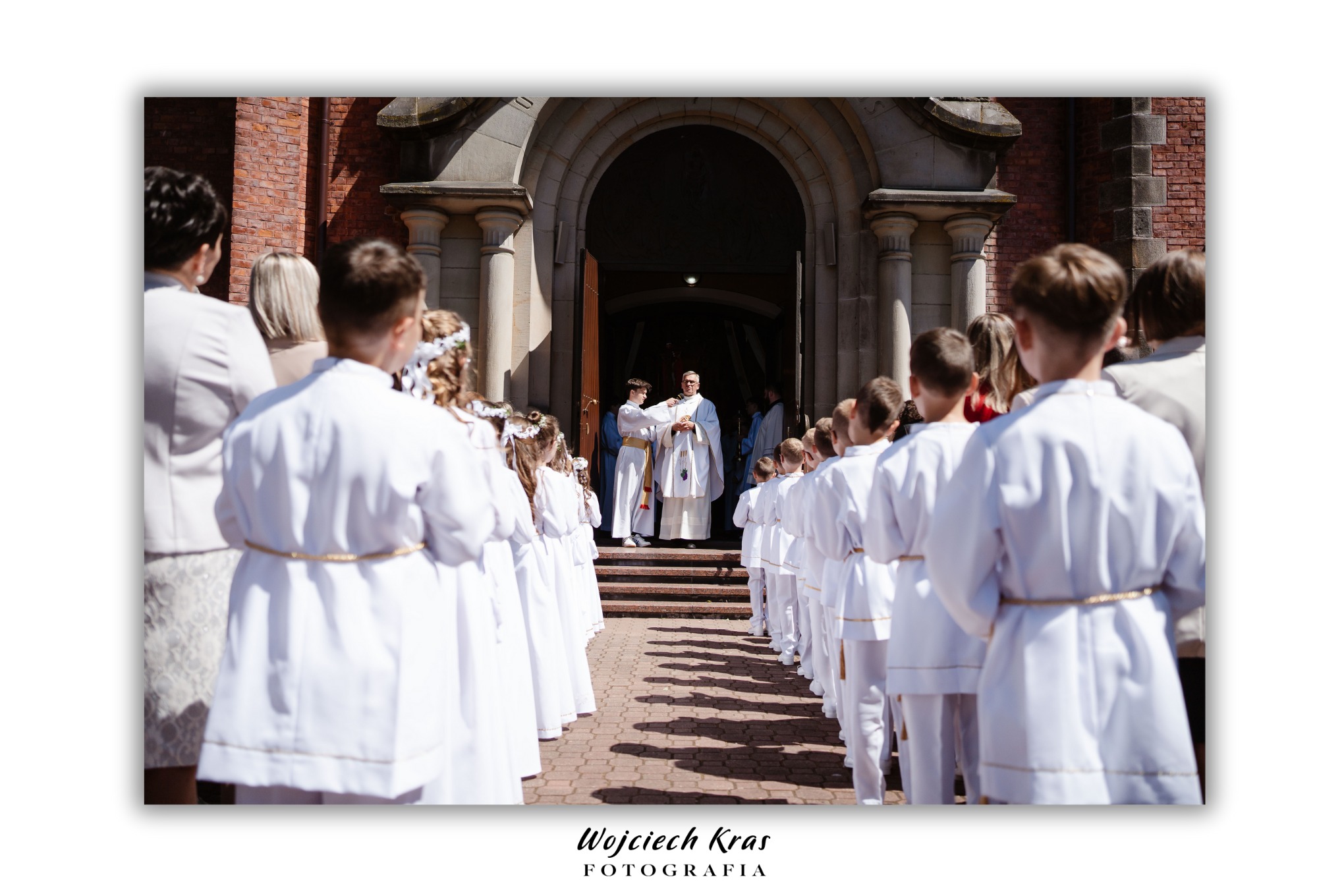 Procesja dzieci w białych strojach komunijnych wchodzących do kościoła, prowadzi ksiądz w ornacie, widok z tyłu na dzieci i rodziców.