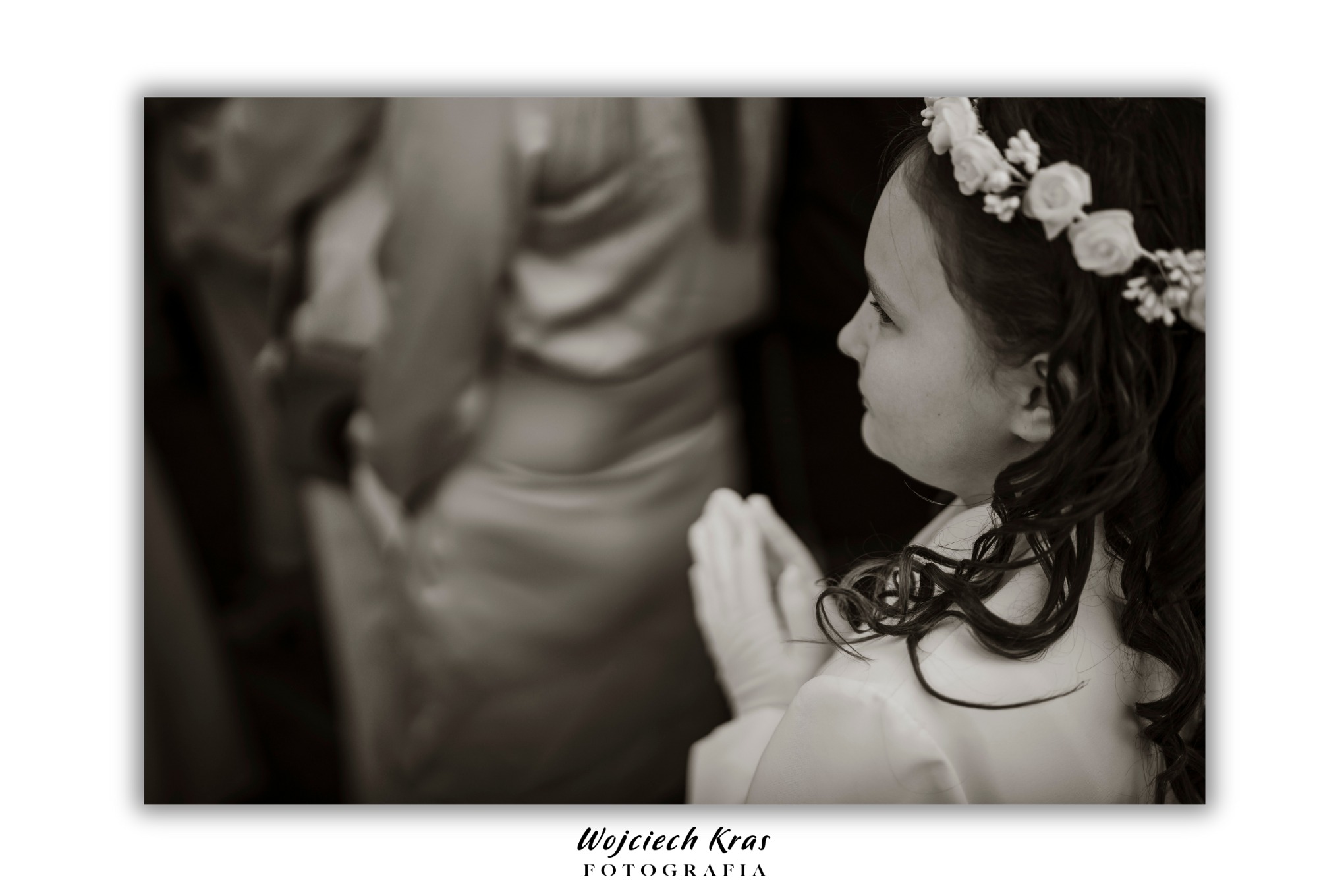 Czarno-białe ujęcie z profilu dziewczynki z wiankiem na głowie, złożone dłonie w białych rękawiczkach, rozmyte tło sukienki