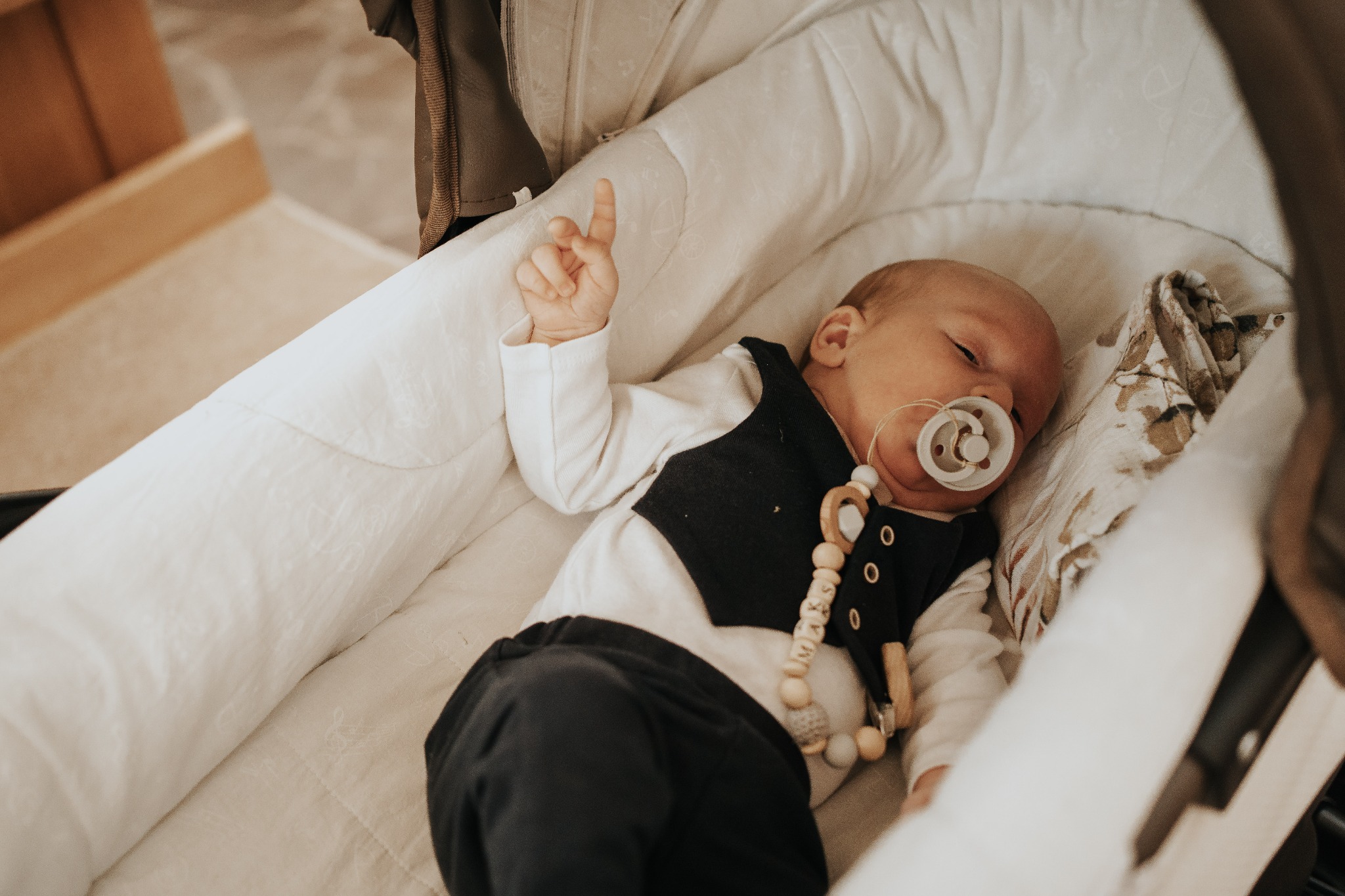 Noworodek śpiący w białym, miękkim koszu Mojżesza z podniesioną rączką, ubrany w białe body, czarną kamizelkę i spodnie, z drewnianym smoczkiem z napisem 'MAKS'.
