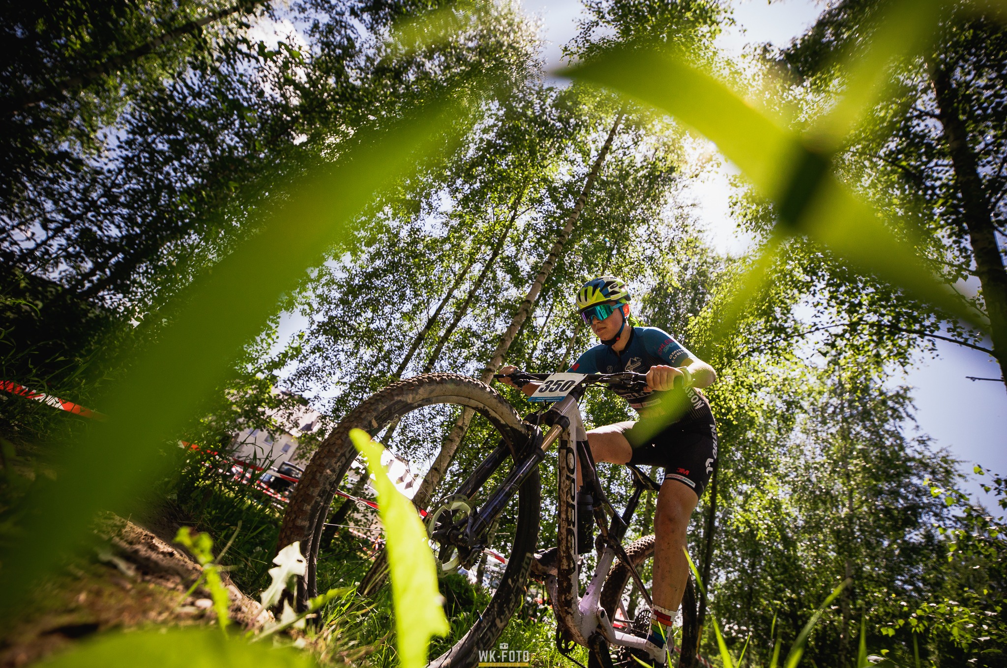 Kolarz górski z numerem 350 na rowerze, jadący leśną ścieżką pod słońce, widok od dołu przez liście.