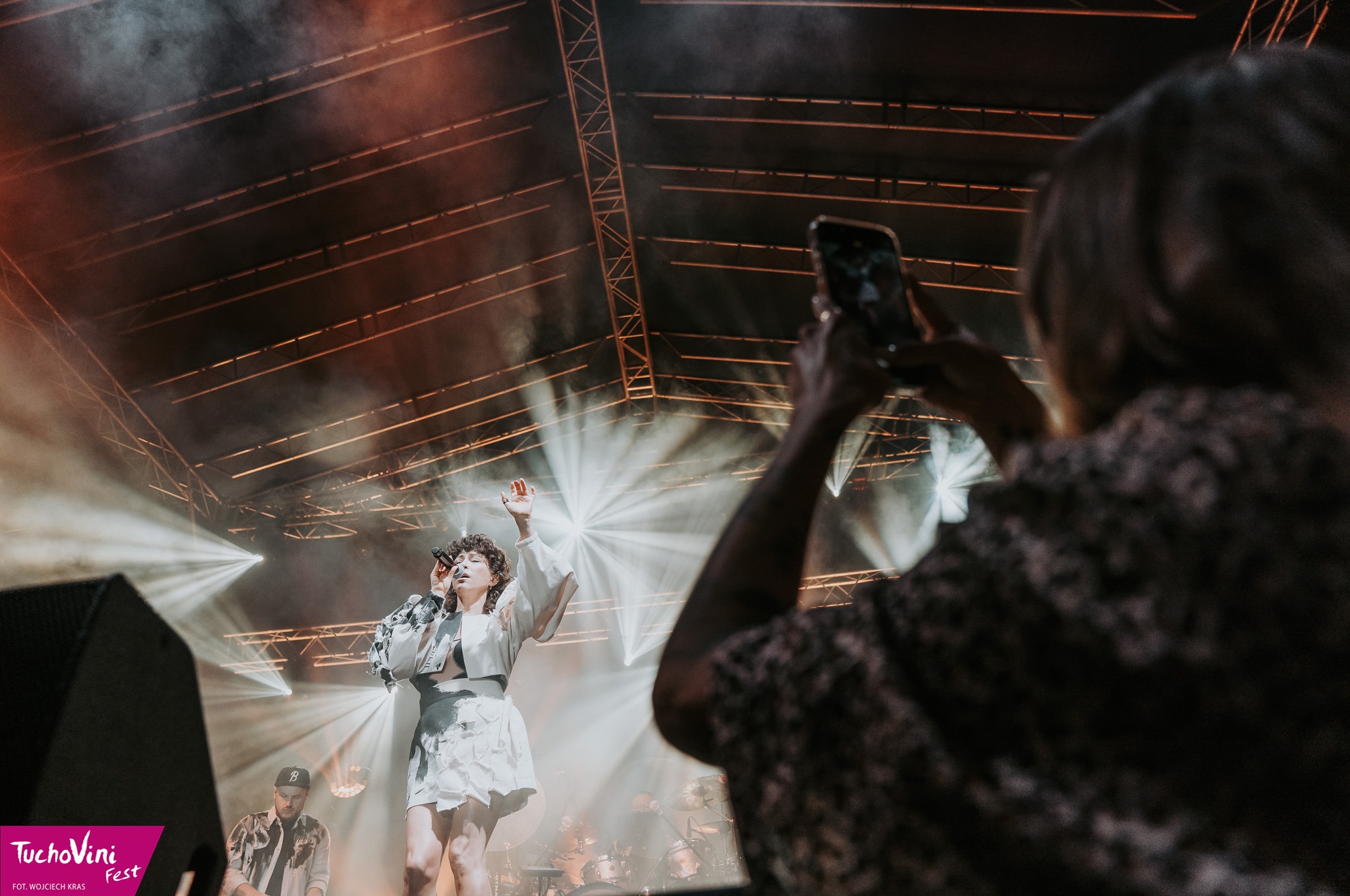 Piosenkarka w białym stroju na scenie podczas koncertu, oświetlona reflektorami, z widoczną publicznością nagrywającą występ telefonem.