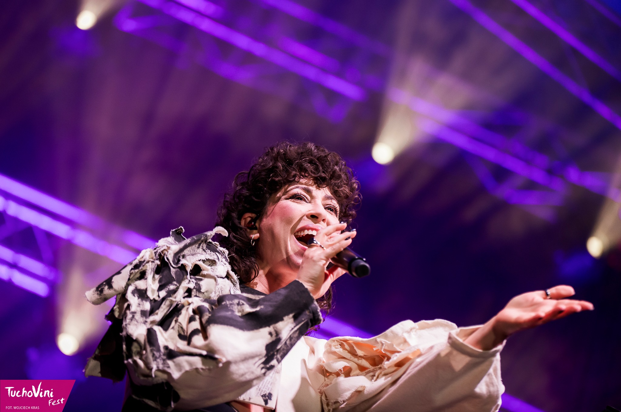 Wokalistka z kręconymi włosami śpiewa do mikrofonu na scenie oświetlonej fioletowymi reflektorami, ubrana w ekstrawagancką kurtkę z wzorem przypominającym krowie łaty.