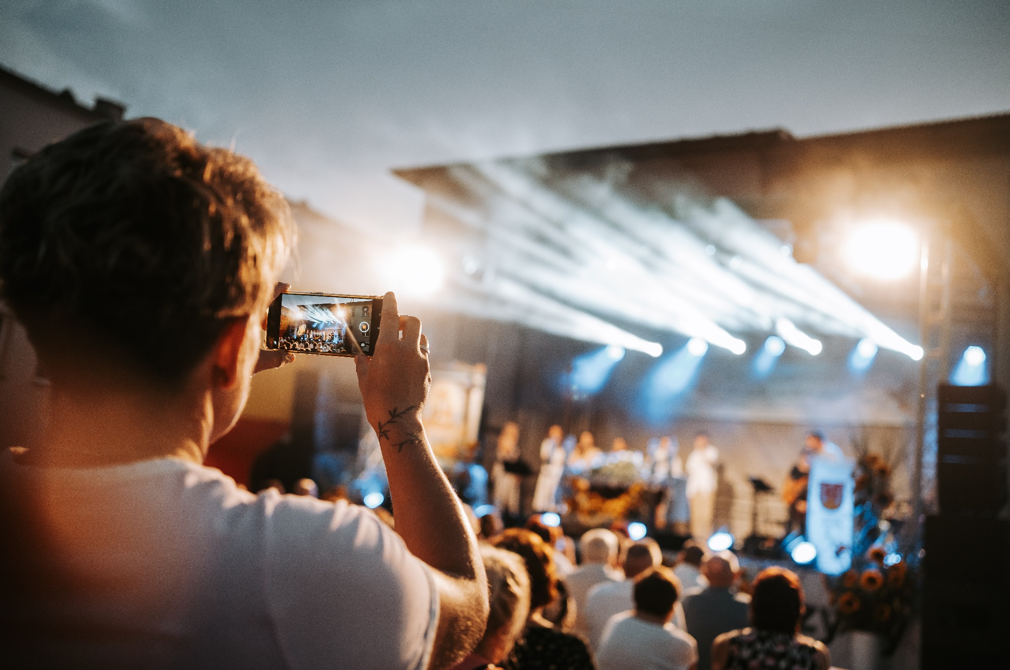 Osoba z tatuażem na przedramieniu filmuje smartfonem koncert na scenie oświetlonej reflektorami, widok zza pleców.