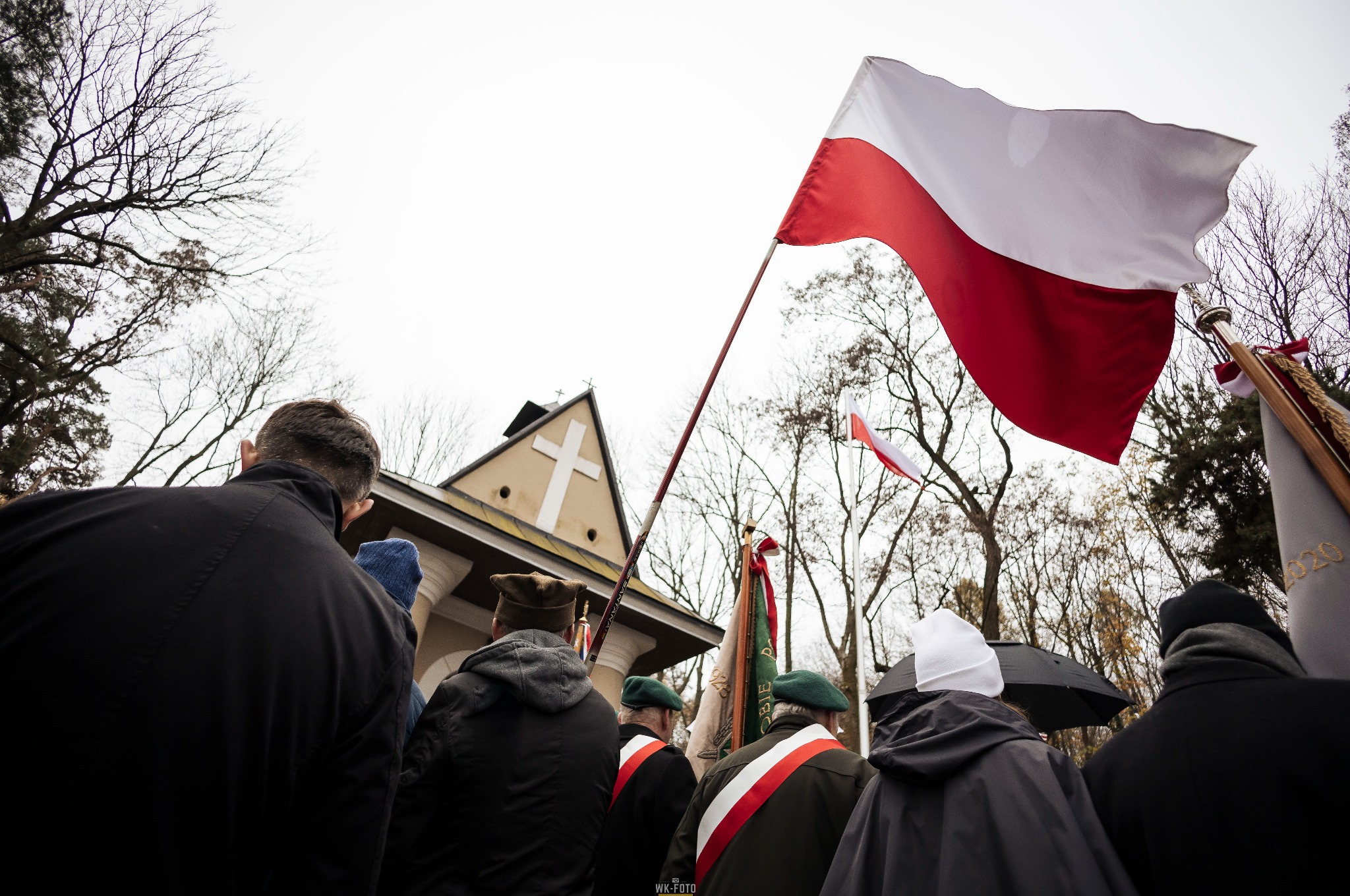 Grupa osób w mundurach z flagą Polski przed budynkiem z krzyżem, pochmurne niebo w tle.