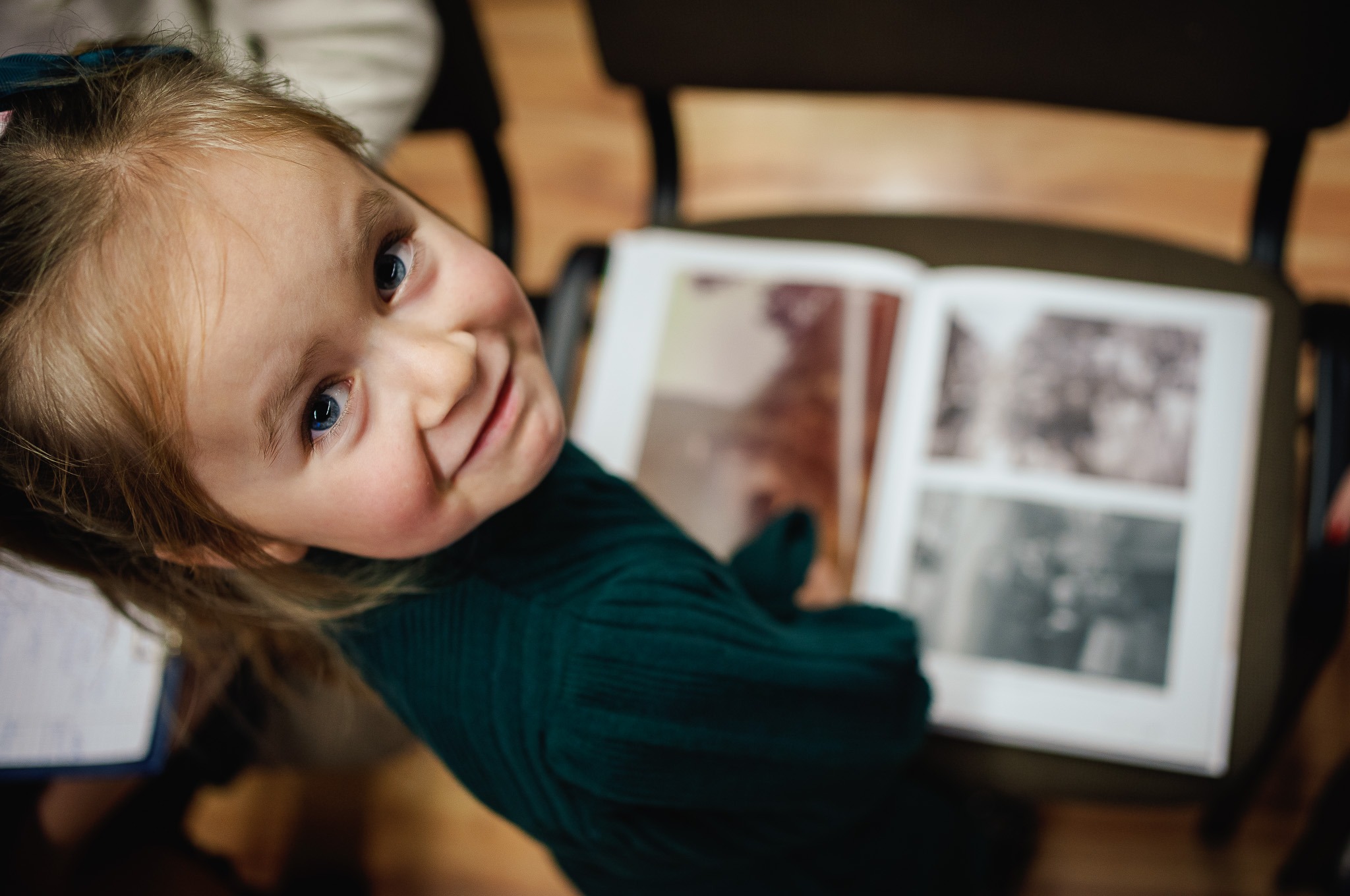 Uśmiechnięta dziewczynka z niebieskimi oczami spogląda w górę, obok otwarty album ze zdjęciami w sepii leżący na krześle.