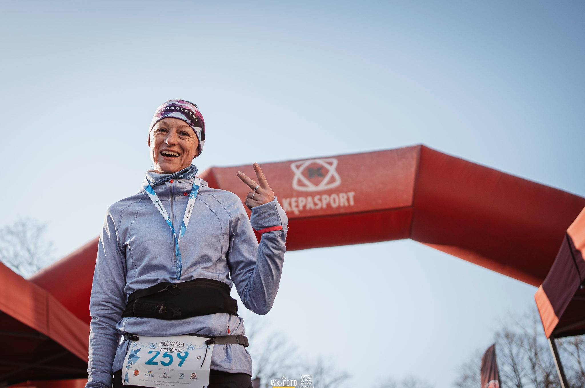 Uśmiechnięta kobieta z numerem startowym 259 na tle czerwonego łuku mety, pokazująca znak zwycięstwa