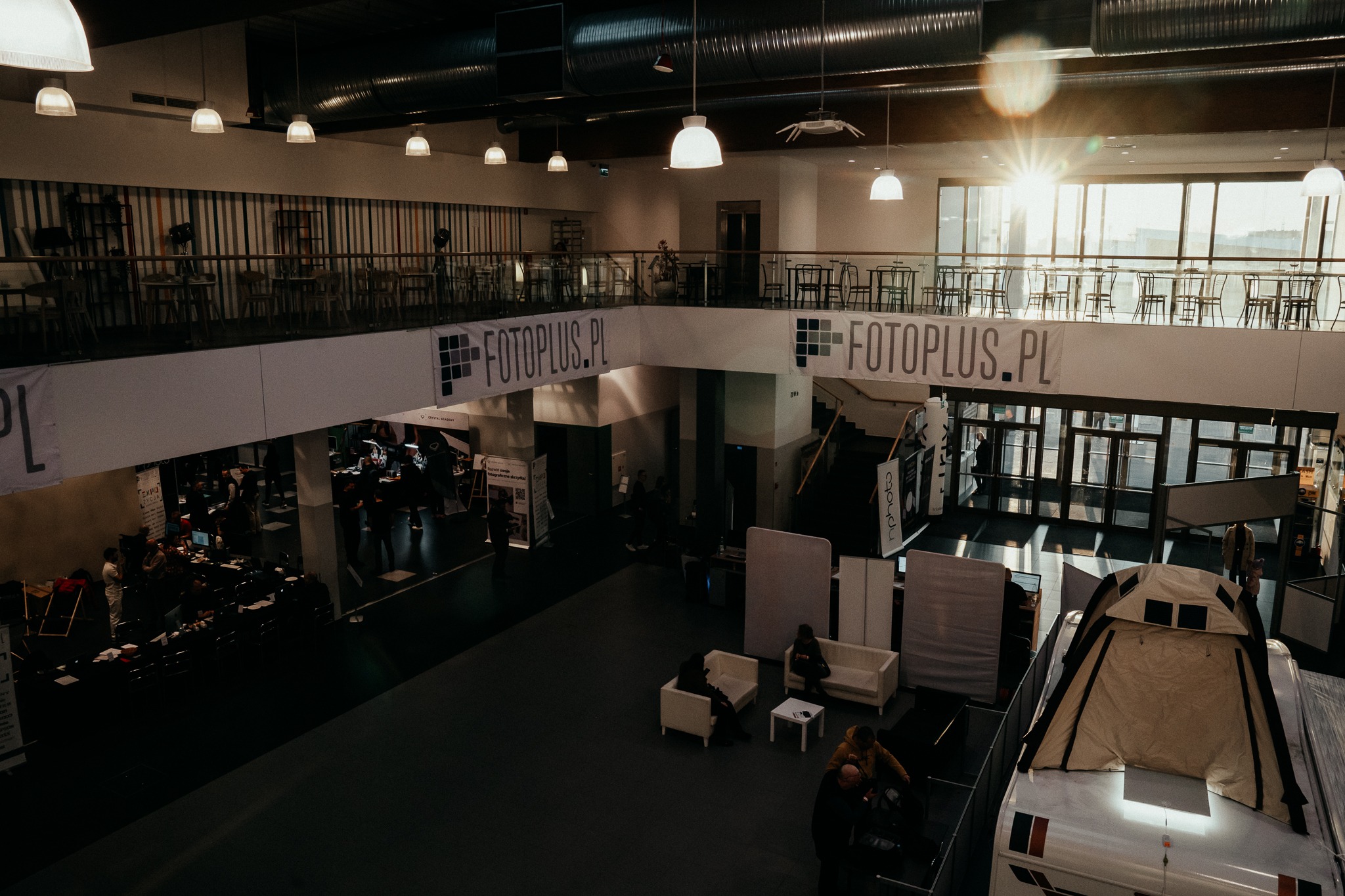 Wnętrze hali wystawienniczej z widocznymi stoiskami, banerami Fotoplus.pl, oświetleniem sufitowym i wpadającym światłem słonecznym przez okna.