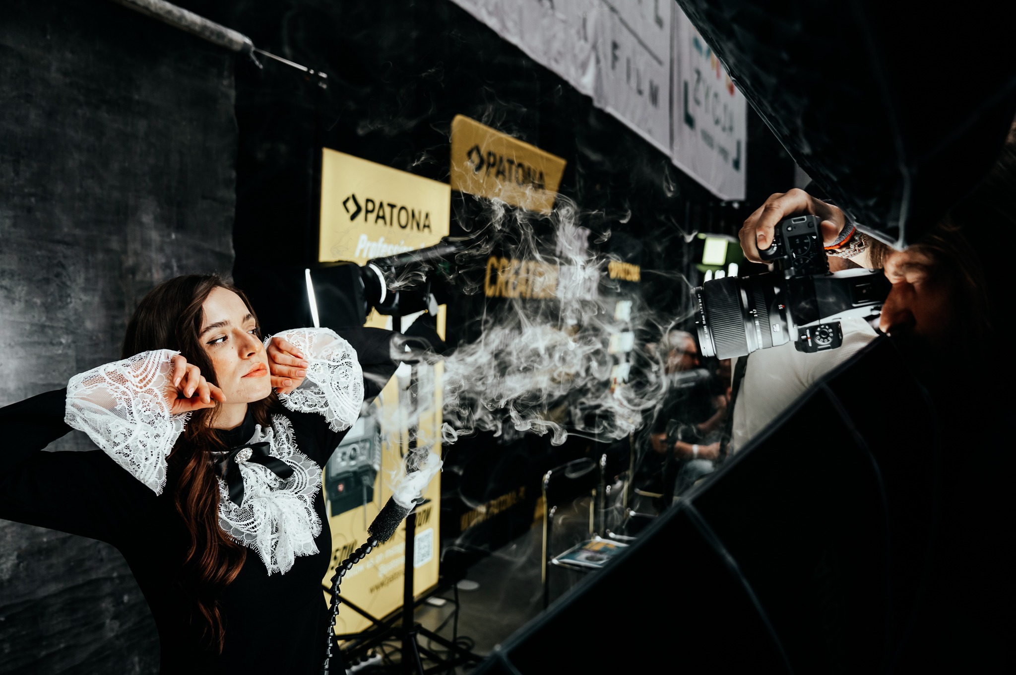 Sesja zdjęciowa w studio: modelka w czarnej sukience z koronkowymi rękawami pozuje na tle studia z dymem, fotograf robi zdjęcie z odbicia w lustrze.