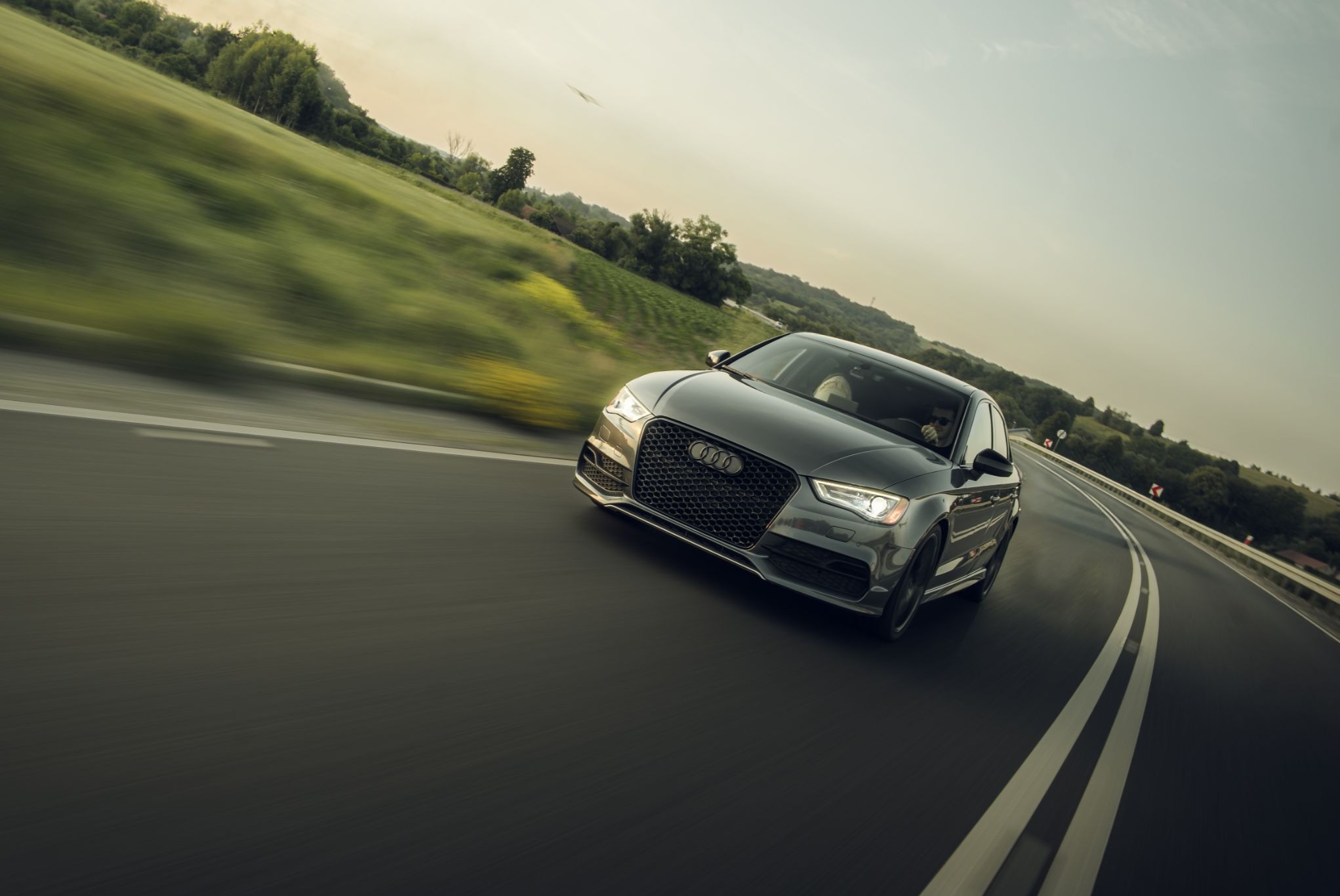 Dynamiczne ujęcie szarego Audi A3 na zakręcie drogi, z rozmytym tłem zielonych wzgórz i pochmurnego nieba, podkreślające prędkość i nowoczesny design auta.