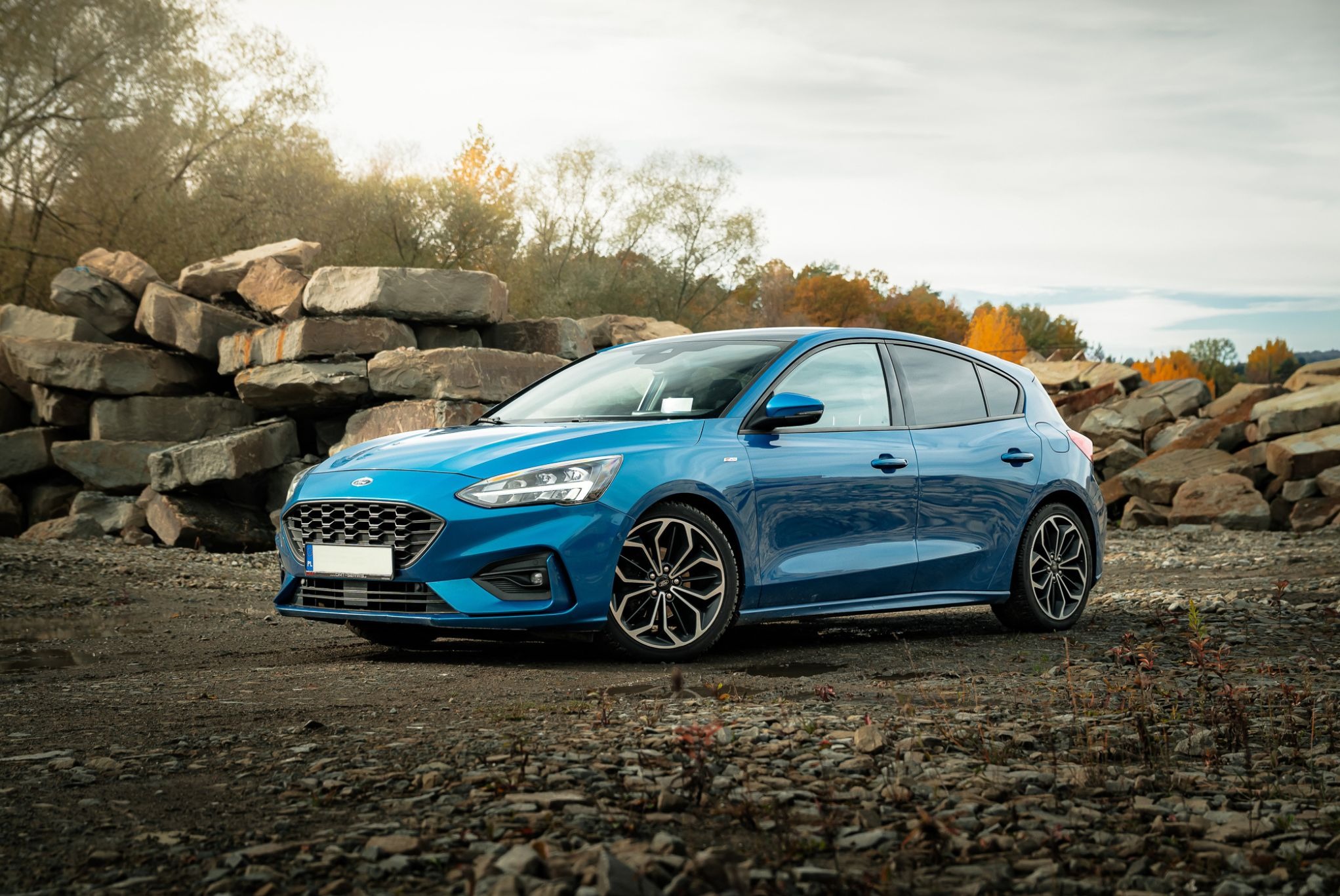 Błękitny Ford Focus hatchback na tle kamiennego nasypu i drzew. Auto ustawione pod kątem, z widocznymi detalami karoserii i felg. Jesienne światło.