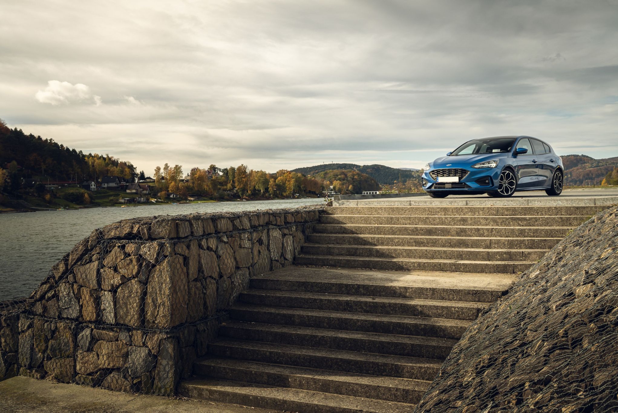 Błękitny Ford Focus na tle jeziora i pochmurnego nieba, ustawiony na betonowych schodach z kamiennym murem. Ujęcie z dołu, podkreślające sylwetkę auta.