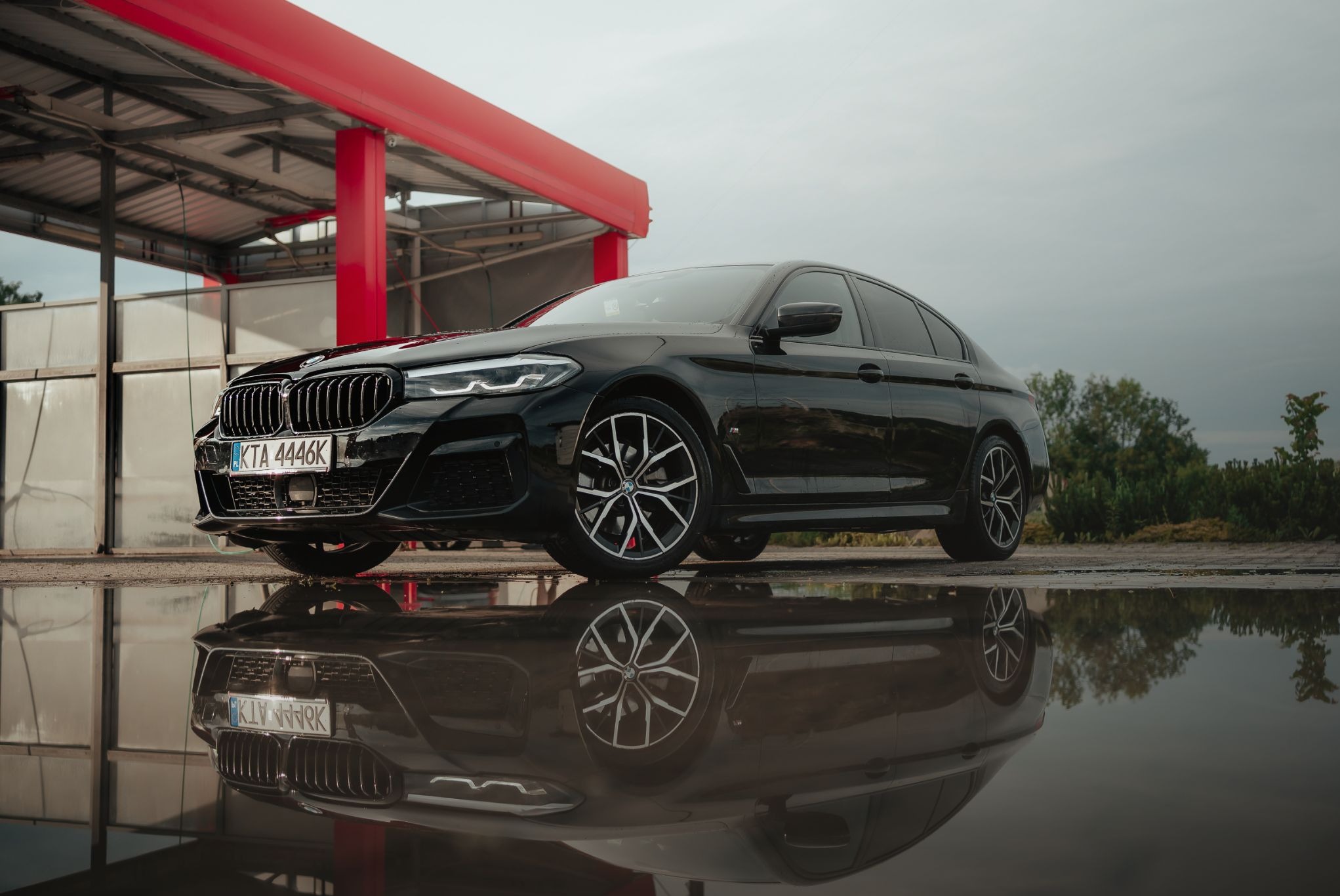 Czarne BMW serii 5 po myciu, odbijające się w kałuży. Widoczny fragment myjni z czerwonymi elementami konstrukcyjnymi. Tablica rejestracyjna KTA 4446K.