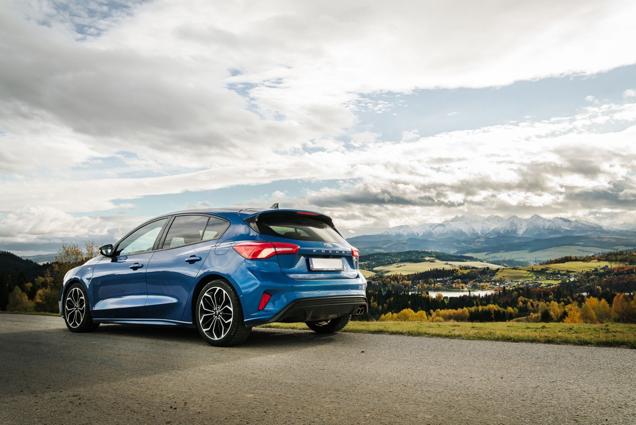 Niebieski Ford Focus hatchback na tle górskiego krajobrazu z jesiennymi drzewami i pochmurnym niebem. Ujęcie z tyłu, podkreślające detale karoserii i felgi.