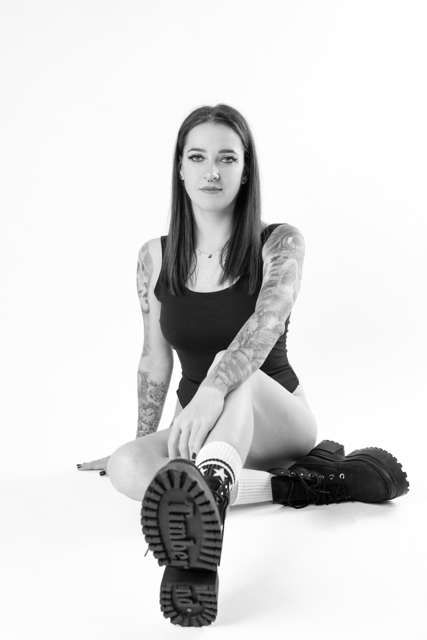 Czarno-białe zdjęcie kobiety siedzącej na podłodze w czarnym body i grubych butach z podeszwą na platformie, eksponującej tatuaże na ramionach i nogach.