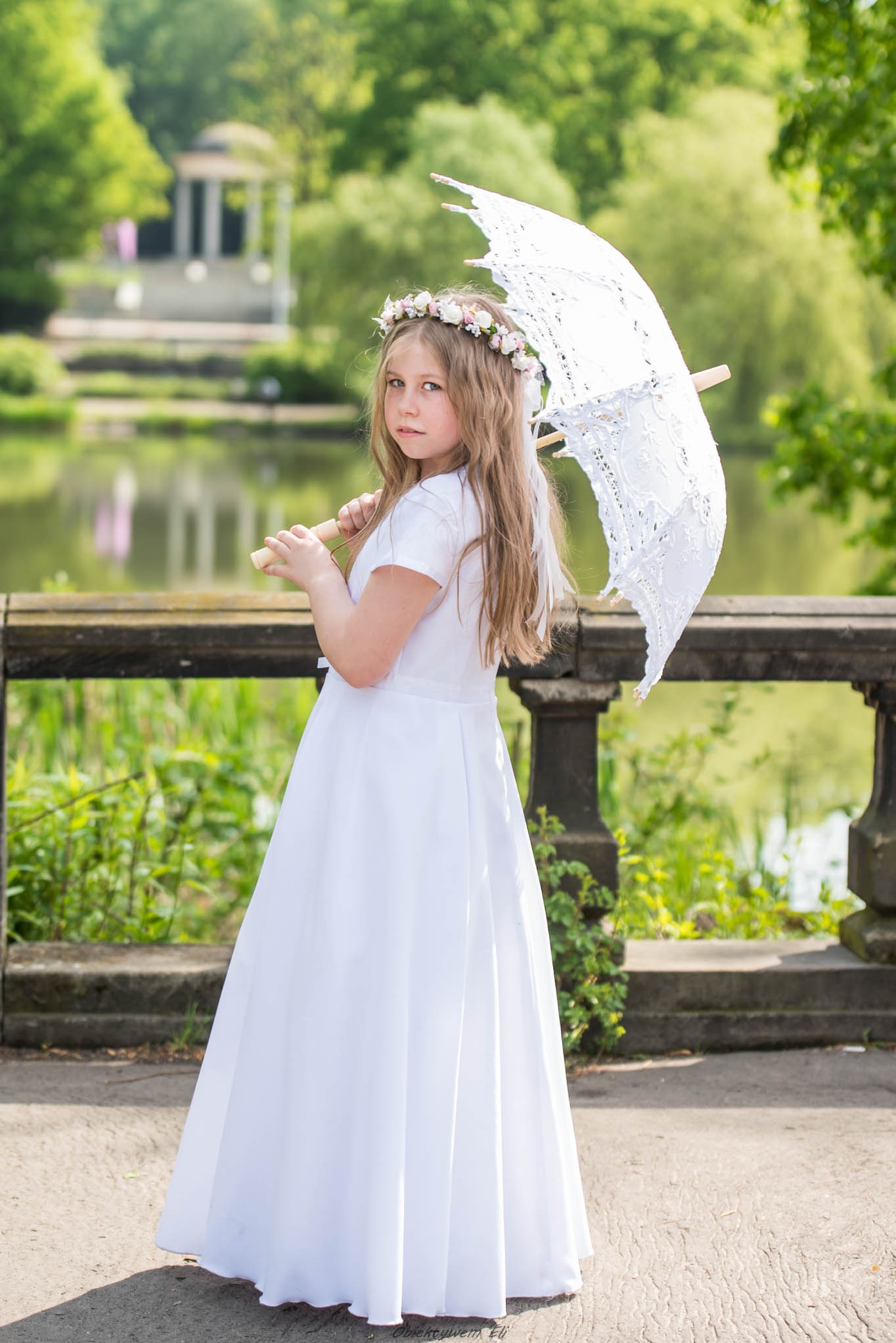 Dziewczynka w białej sukience komunijnej, z wiankiem na głowie i koronkowym parasolem, pozuje na tle parku z jeziorem i altaną.