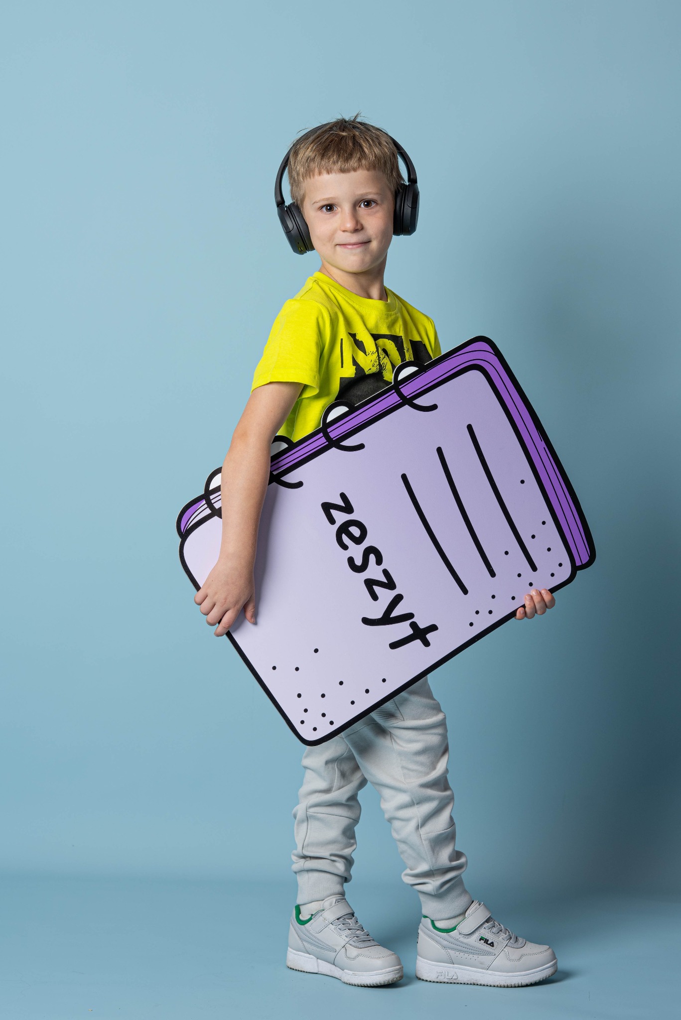 Uśmiechnięty chłopiec w słuchawkach trzyma duży, rysunkowy model zeszytu z napisem 'zeszyt' na jasnoniebieskim tle.
