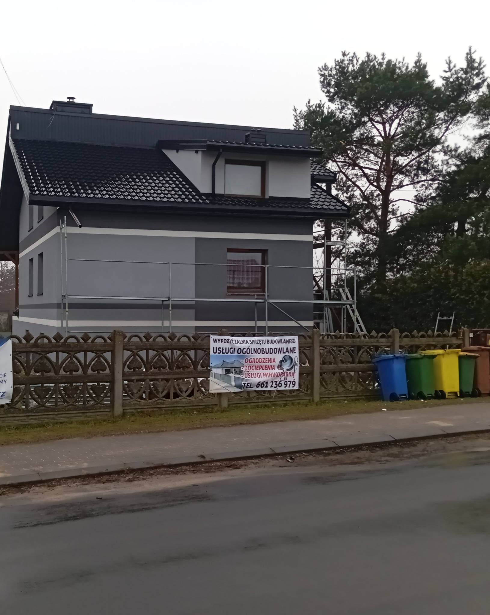 Dom jednorodzinny w trakcie ocieplania, widoczne rusztowania, szary odcień elewacji, w tle drzewa iglaste, przed posesją ozdobny betonowy płot i kolorowe pojemniki na śmieci, na płocie baner...