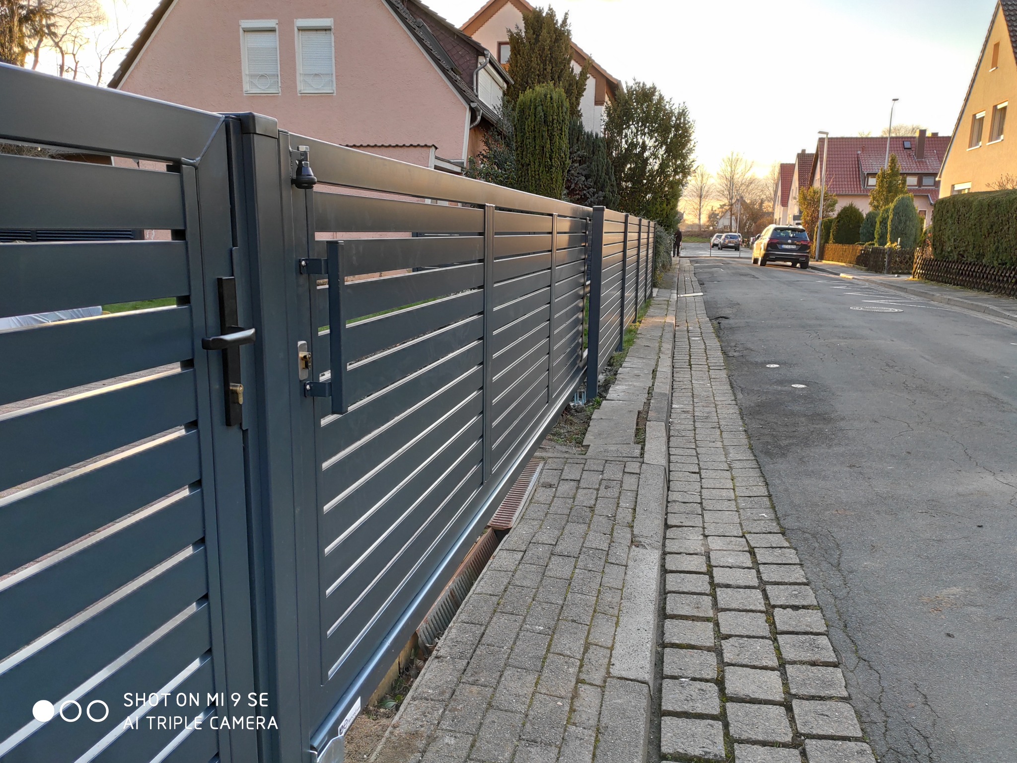 Długa, szara przesuwana brama z poziomymi panelami na tle ulicy i domów jednorodzinnych.