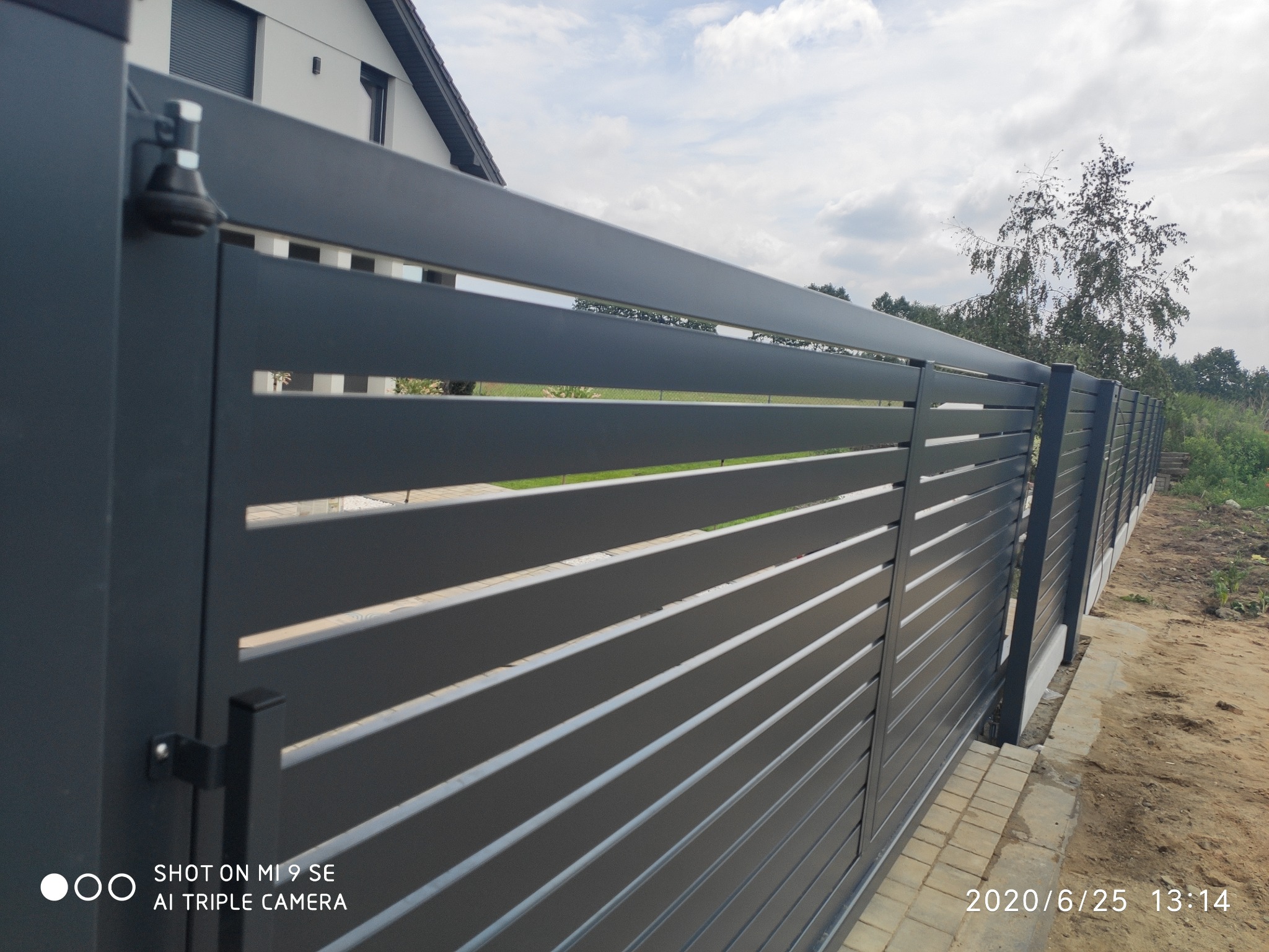 Nowoczesna, pozioma brama wjazdowa z szarych, metalowych profili, widoczny zamek i fragment posesji z kostką brukową.