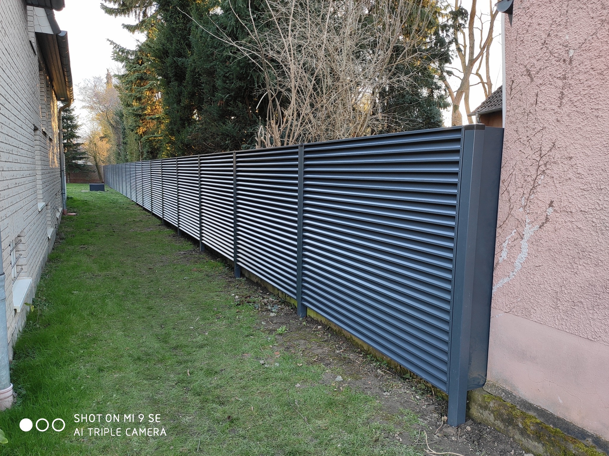 Długa, pozioma, szara przesłona ogrodzeniowa z lamelami, oddzielająca posesje z trawnikiem i drzewami w tle, widok z boku.