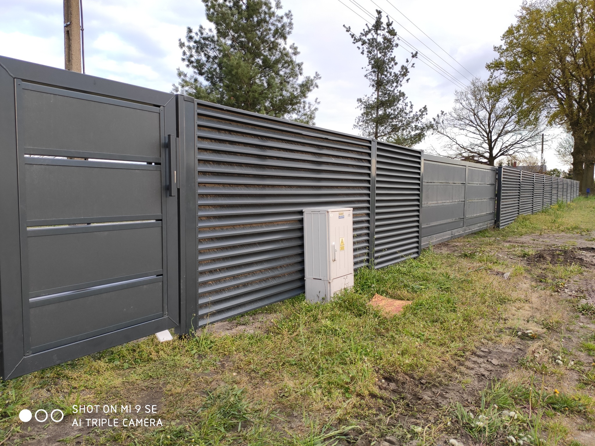 Nowoczesne, szare ogrodzenie z poziomych paneli metalowych i przesuwna brama wjazdowa, widoczna skrzynka elektryczna na trawiastym terenie.