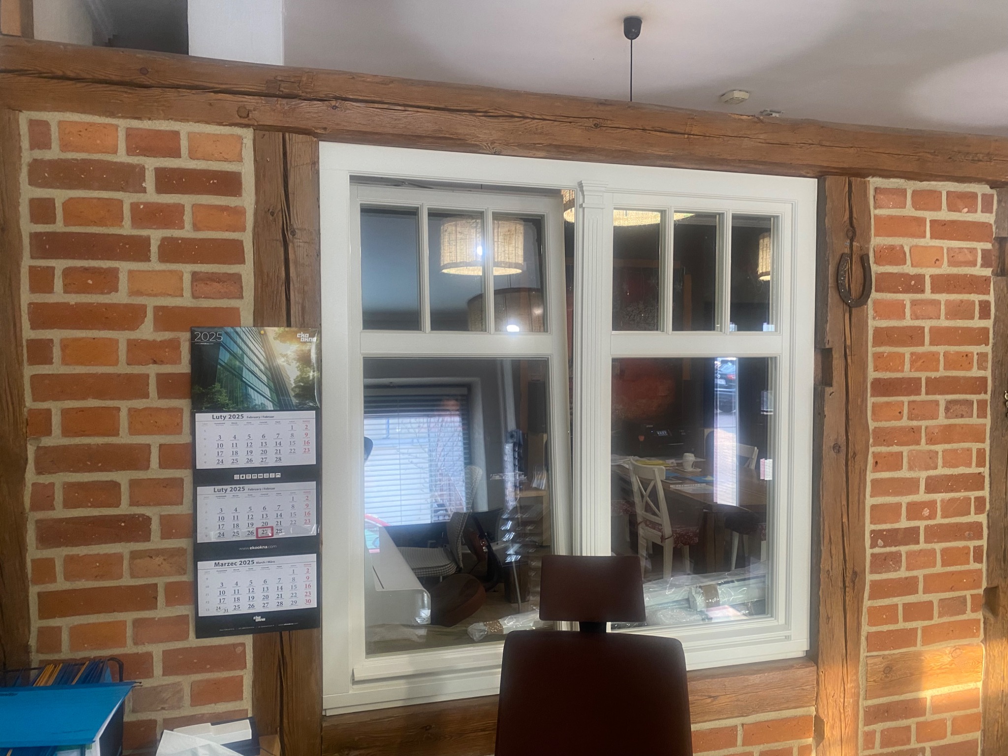 Nowe, białe okno PCV zamontowane w ścianie z czerwonej cegły i drewnianych belkach, widoczny kalendarz na ścianie, wnętrze domu w tle.