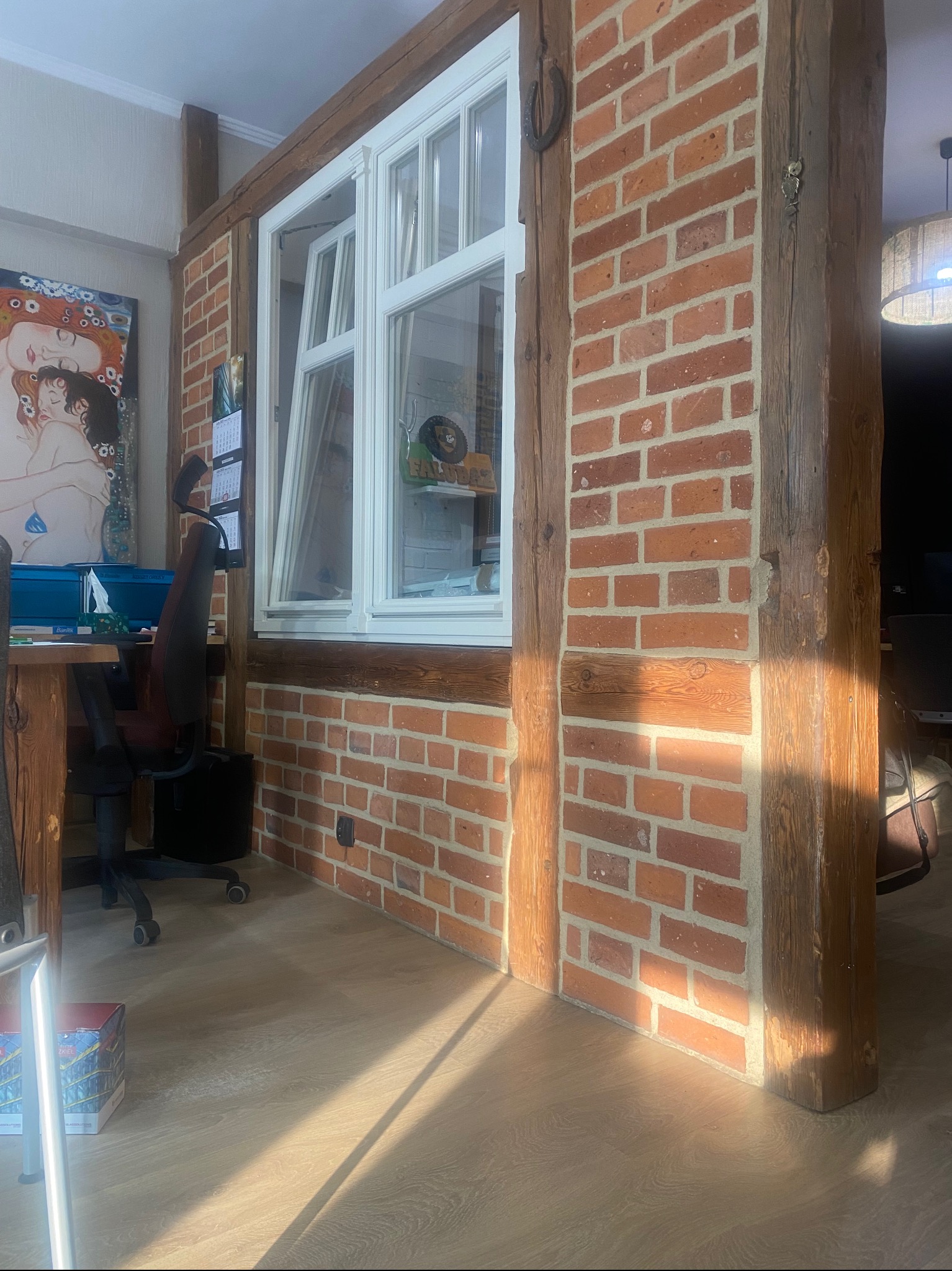 Fragment odnowionego wnętrza w Świebodzinie z ceglaną ścianą i drewnianymi belkami, widoczne biurko i krzesło, na ścianie obraz i kalendarz.