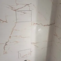 Wykończona ściana łazienki z białych płytek imitujących marmur z złotymi żyłami, widoczne dwa otwory rewizyjne i wystające rury instalacji wodnej.