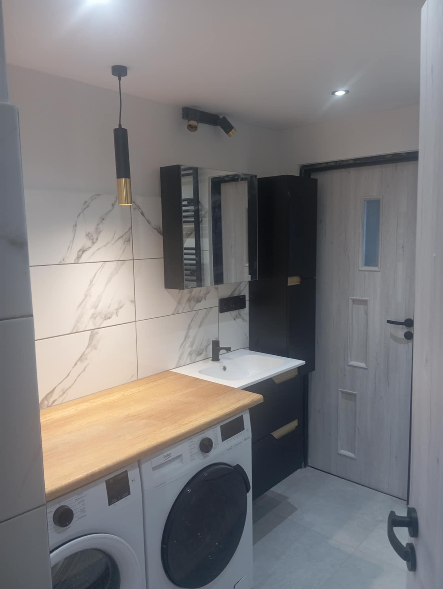 Nowoczesna łazienka z marmurowymi płytkami, drewnianym blatem nad pralką i czarnymi szafkami. Minimalistyczna lampa wisząca i oświetlenie punktowe.