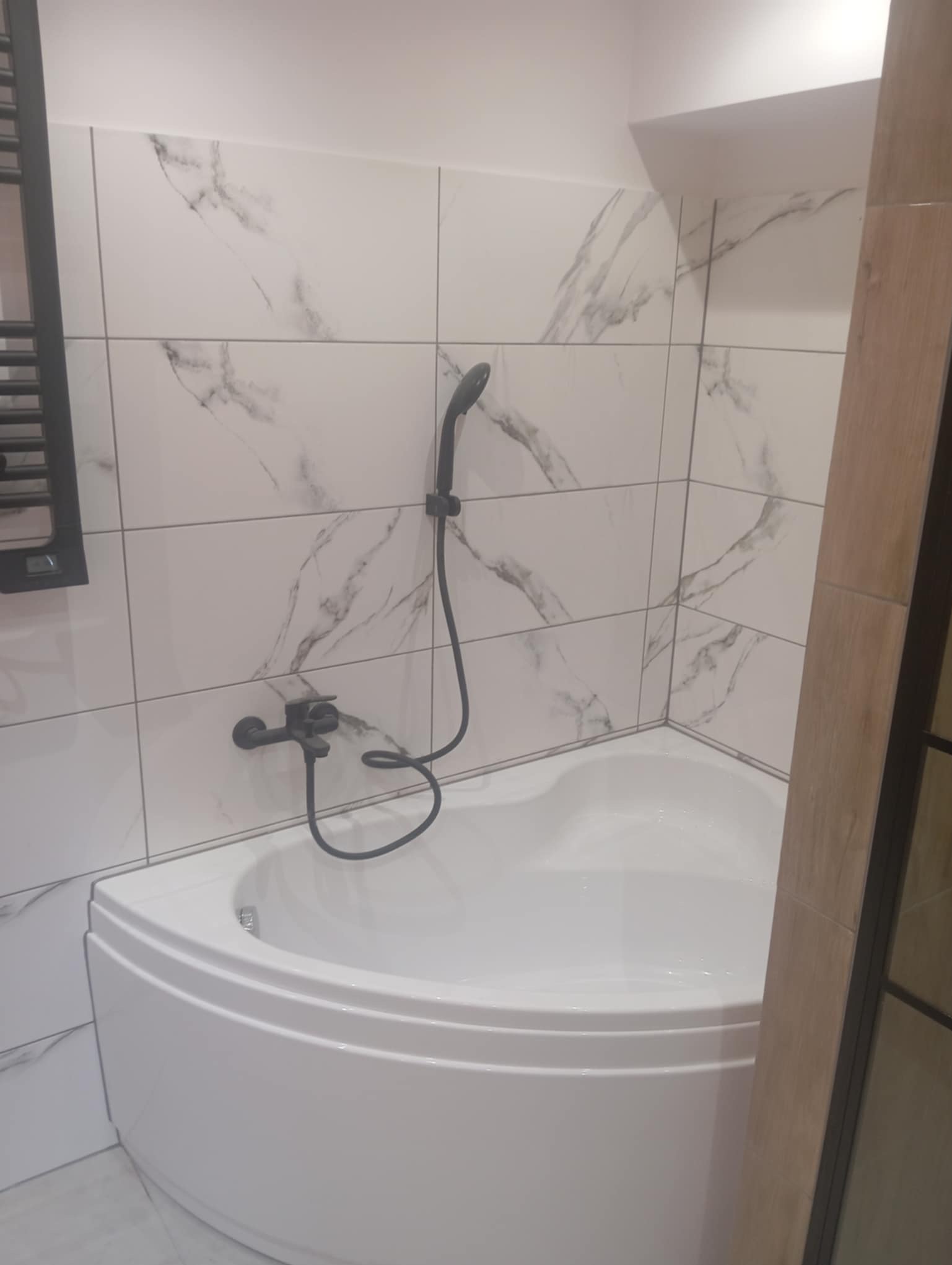 Biała narożna wanna z czarną armaturą i marmurowymi płytkami ściennymi. Nowoczesna łazienka po remoncie, widok na fragment kabiny prysznicowej.