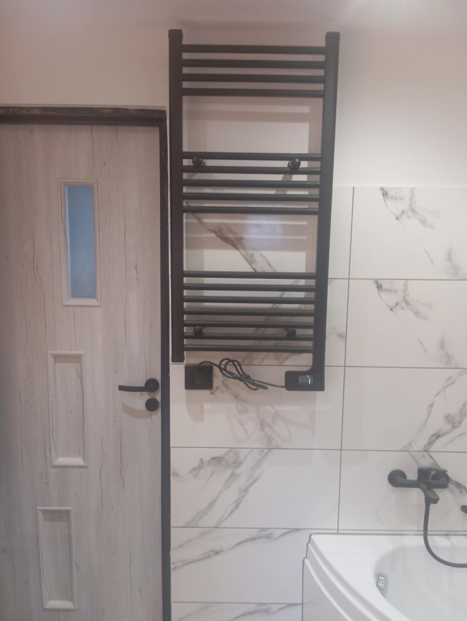 Wykończona łazienka w Świdniku: białe płytki imitujące marmur, czarny grzejnik drabinkowy z elementami elektrycznymi, drzwi z drewnopodobnym wzorem.