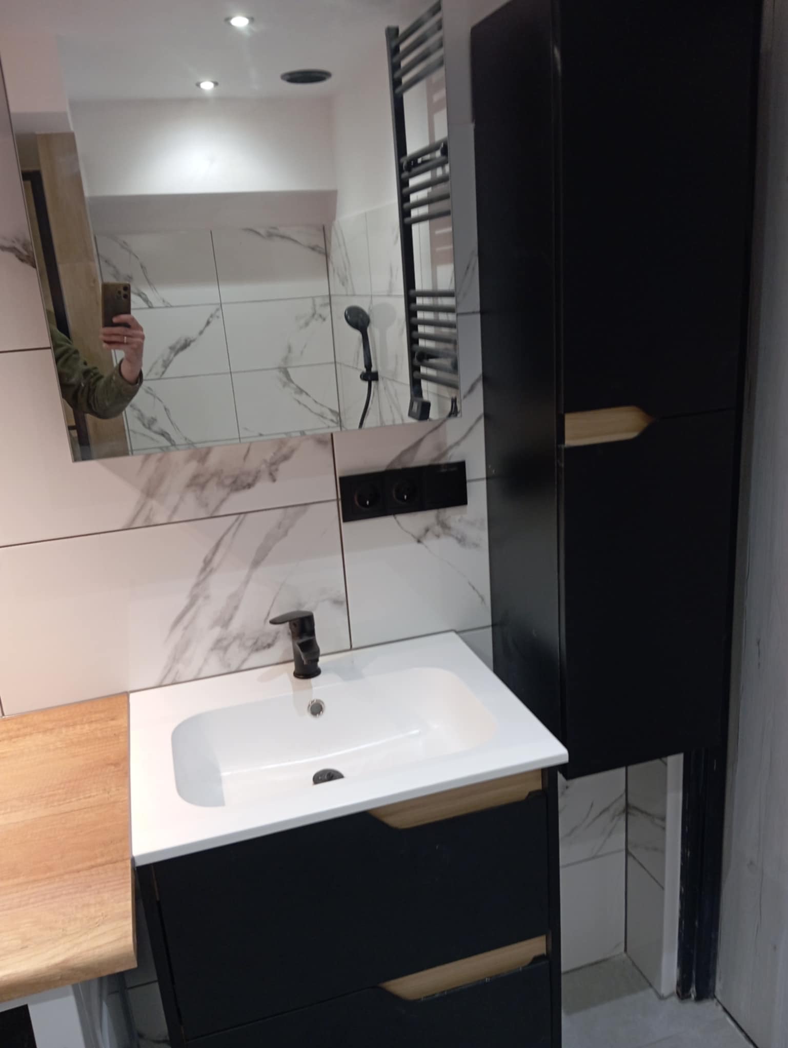 Nowoczesna łazienka po remoncie: białe płytki imitujące marmur, czarna szafka pod umywalką z drewnianymi akcentami, grzejnik drabinkowy i lustro.